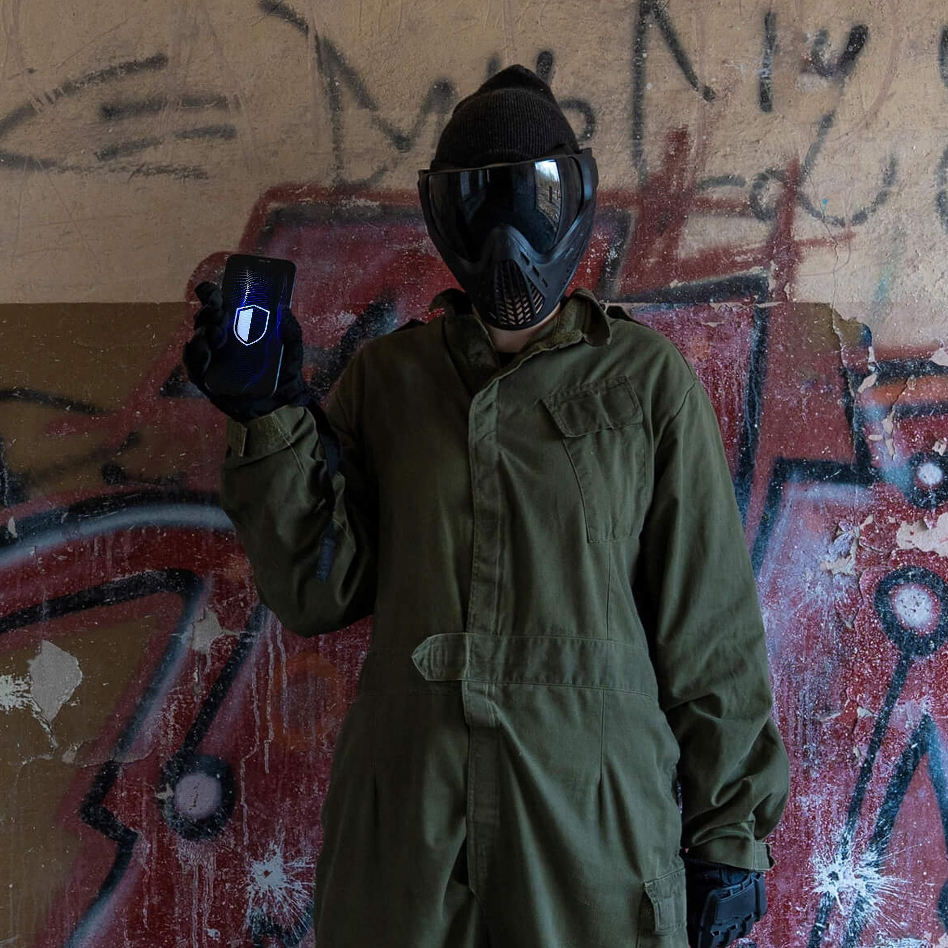 Una persona in tuta verde e maschera tattica mostra uno smartphone con un'icona a scudo davanti a un muro graffitato.