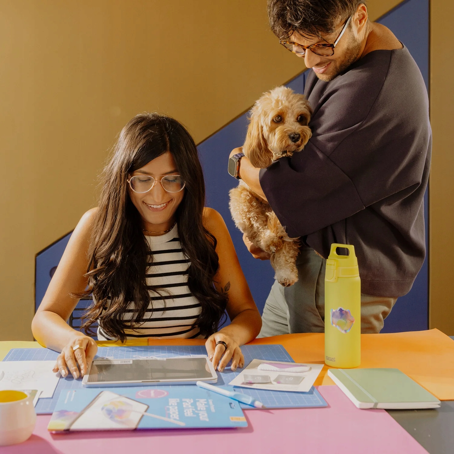 Una donna sorridente usa un tablet su una scrivania colorata mentre un uomo tiene un cagnolino.