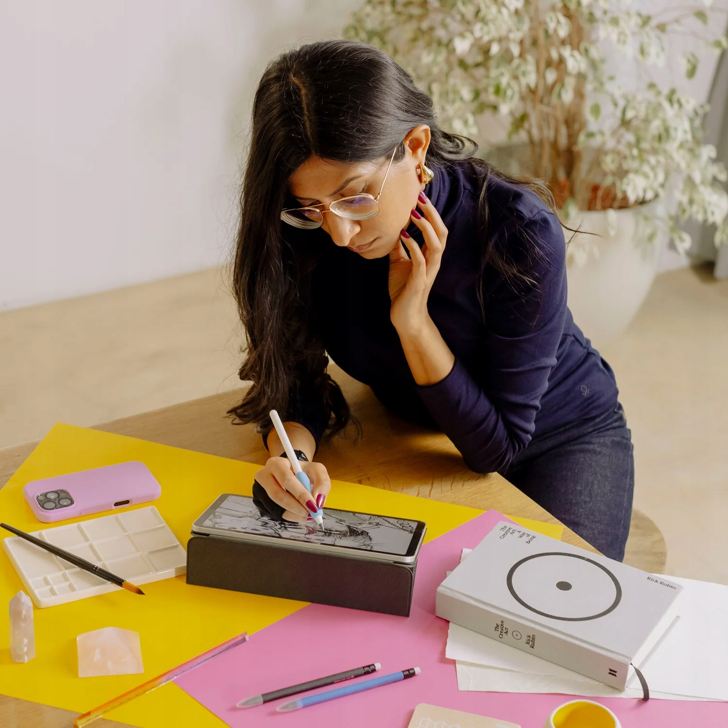 Una donna con un maglione blu disegna con un pennino su un tablet a una scrivania con carte colorate.