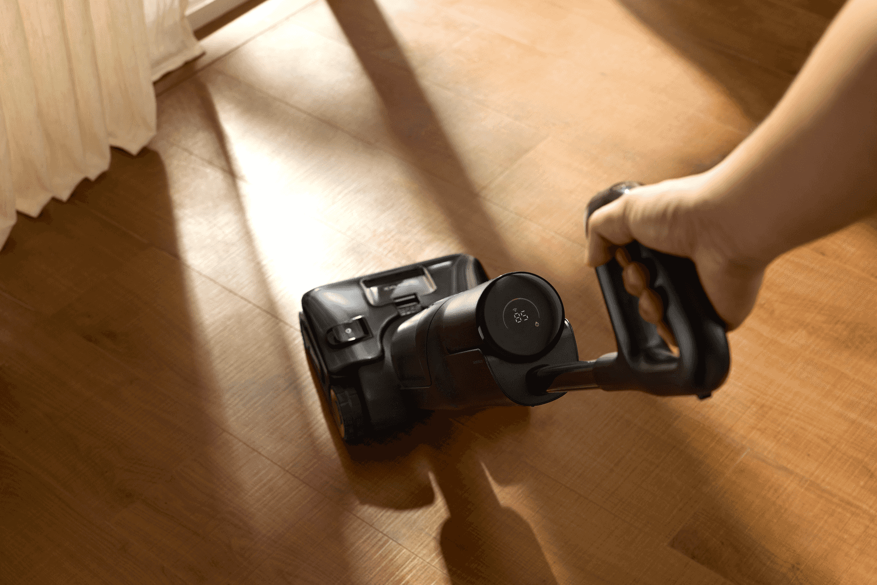 Una mano guía una aspiradora inalámbrica negra sobre un piso de madera en una habitación soleada.