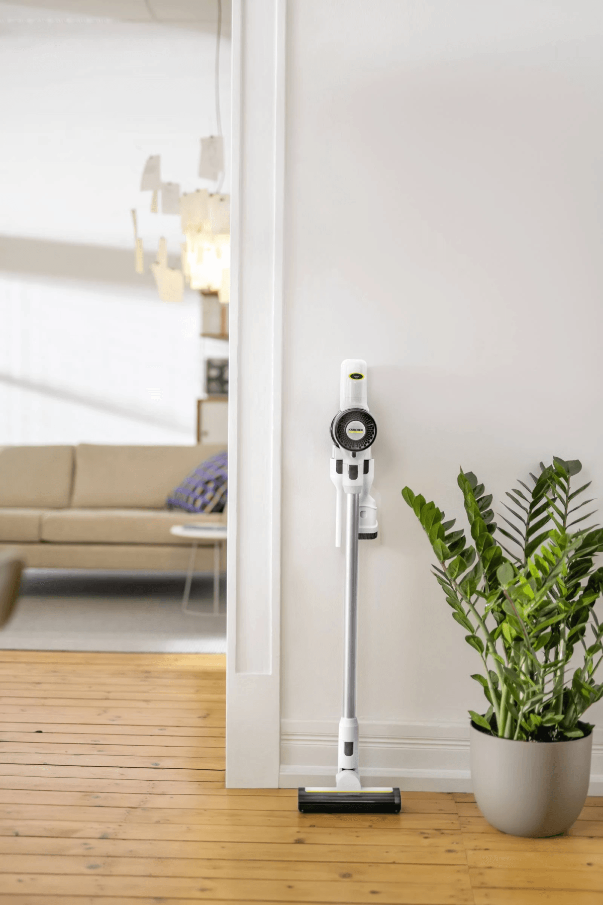 Una aspiradora inalámbrica blanca montada en una pared blanca junto a una planta en maceta, con una sala de estar de fondo.