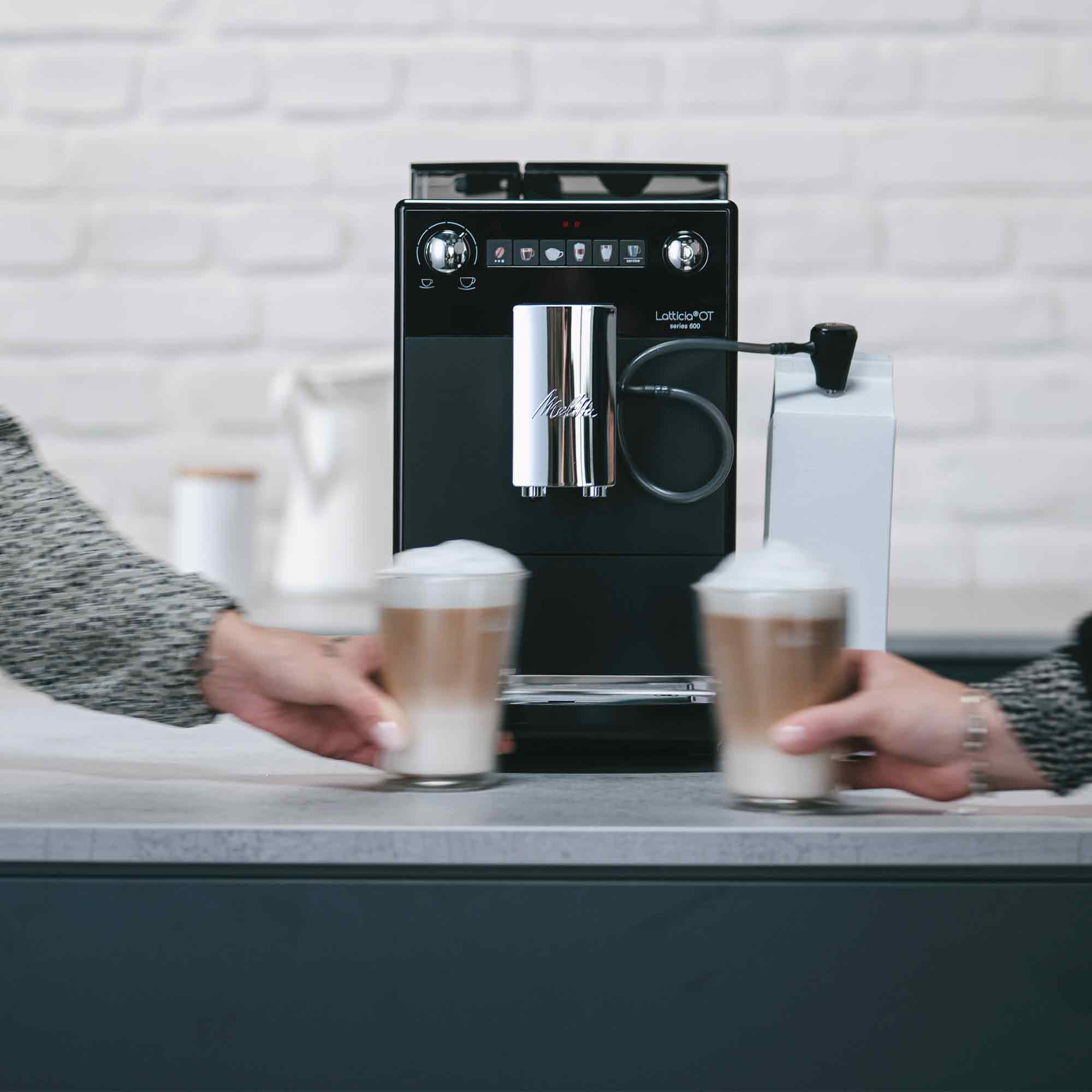 Due persone prendono bicchieri di latte macchiato da una macchina da caffè nera davanti a un muro di mattoni bianchi.