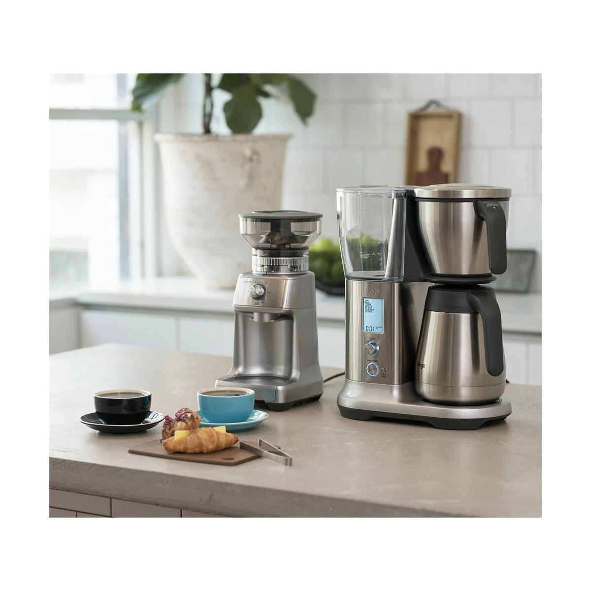 Una macchina da caffè e macinacaffè su un bancone con due tazze di caffè e cornetti in una cucina luminosa.