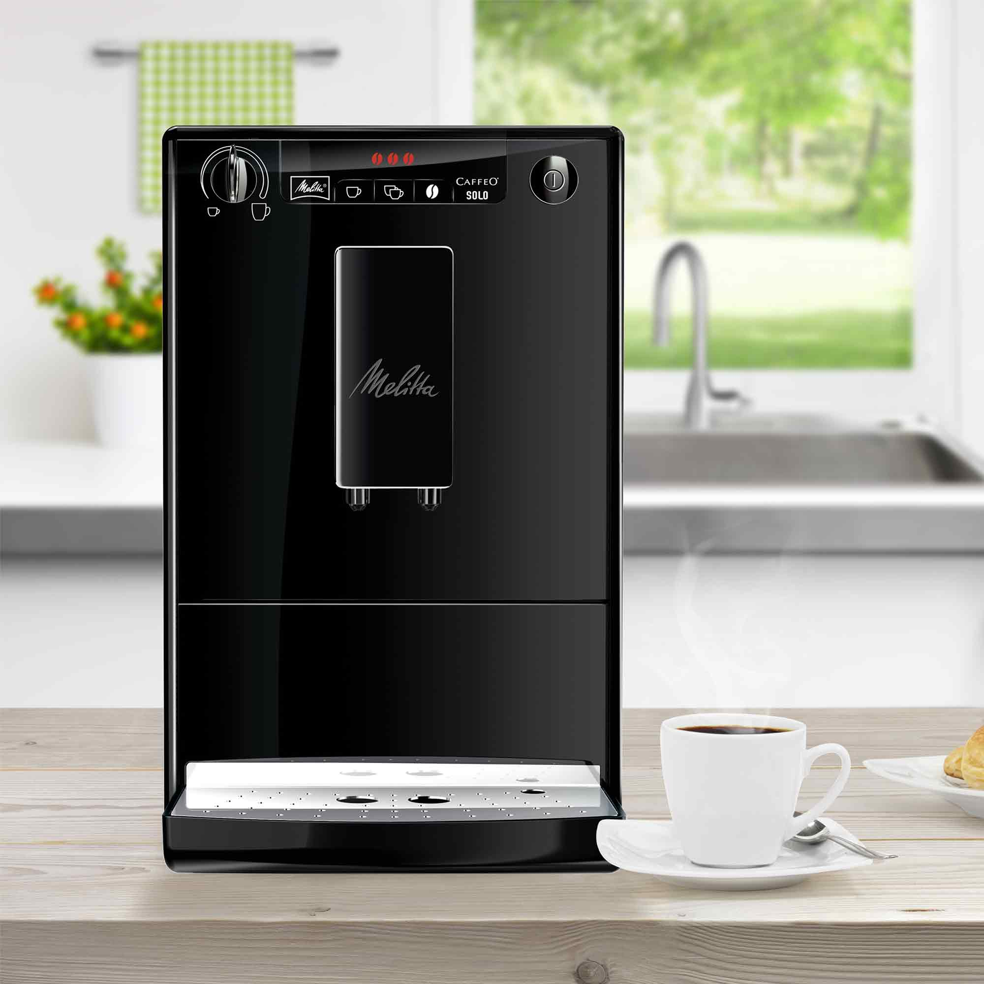 Een zwarte Melitta-koffiemachine staat op een houten tafel naast een kopje verse koffie in een keuken.