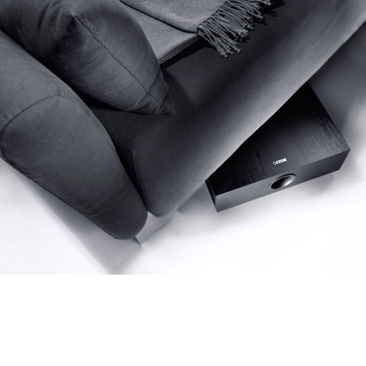 Ein graues Sofa mit einer Decke und Kissen, darunter befindet sich ein schwarzer Lautsprecher.