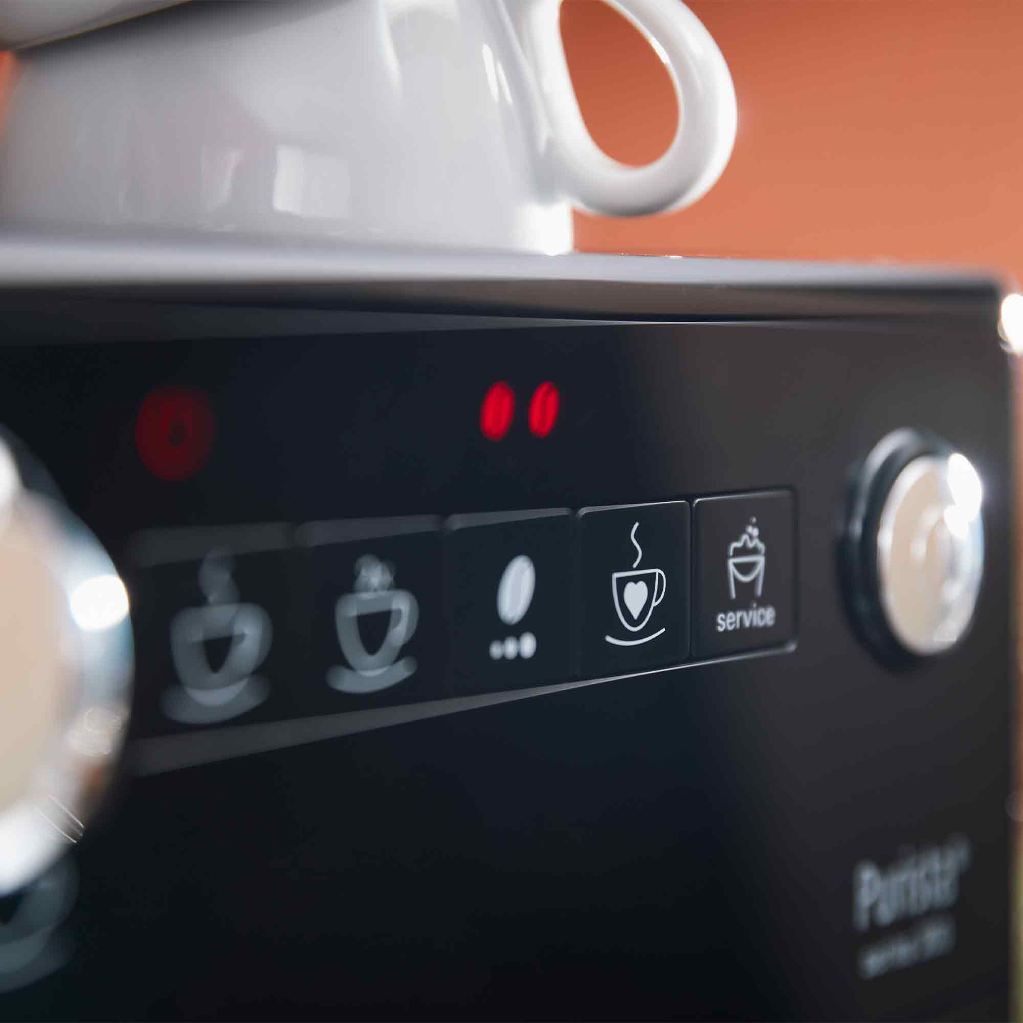 Nahaufnahme des Bedienfelds einer Kaffeemaschine mit beleuchteten Tasten und einer weißen Tasse oben drauf.