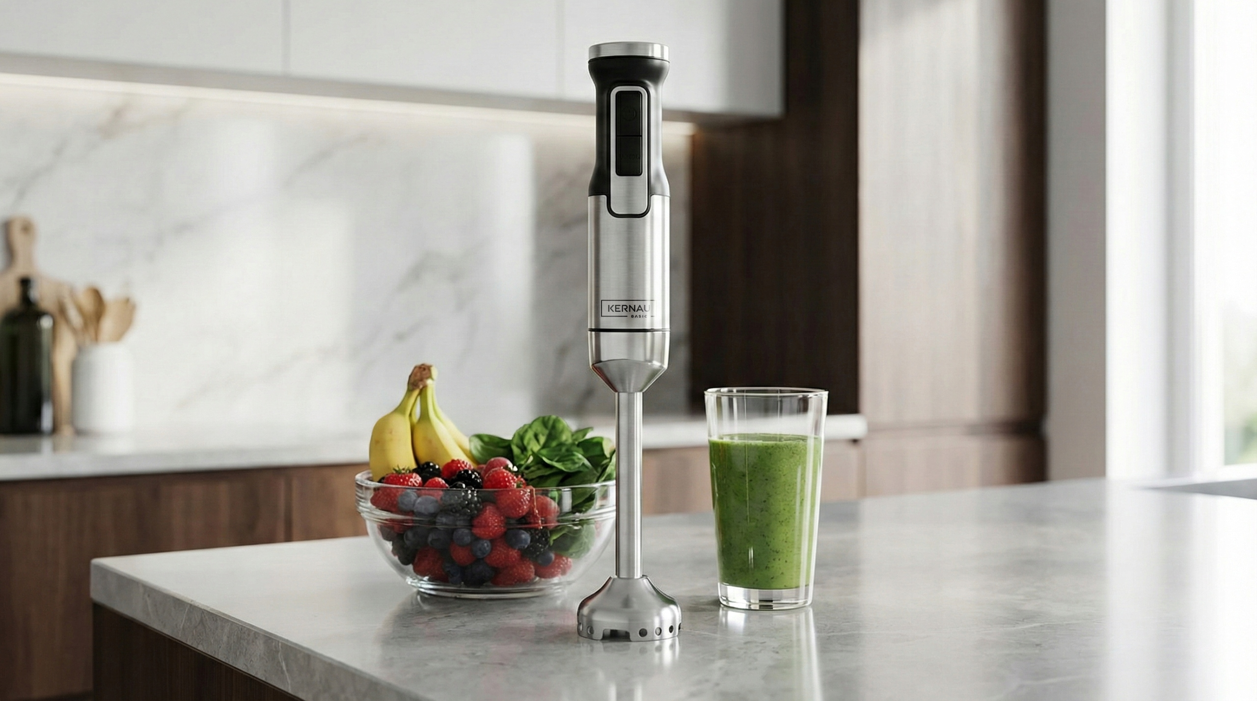 Srebrny blender ręczny stoi na blacie kuchennym obok miski z owocami i szklanki zielonego smoothie.