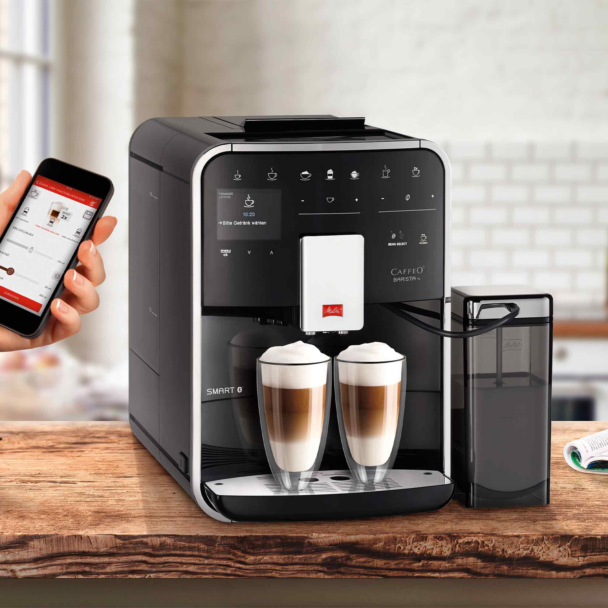 Eine schwarz-silberne Melitta Kaffeemaschine brüht zwei Latte auf einer Holztheke, gesteuert per Smartphone.
