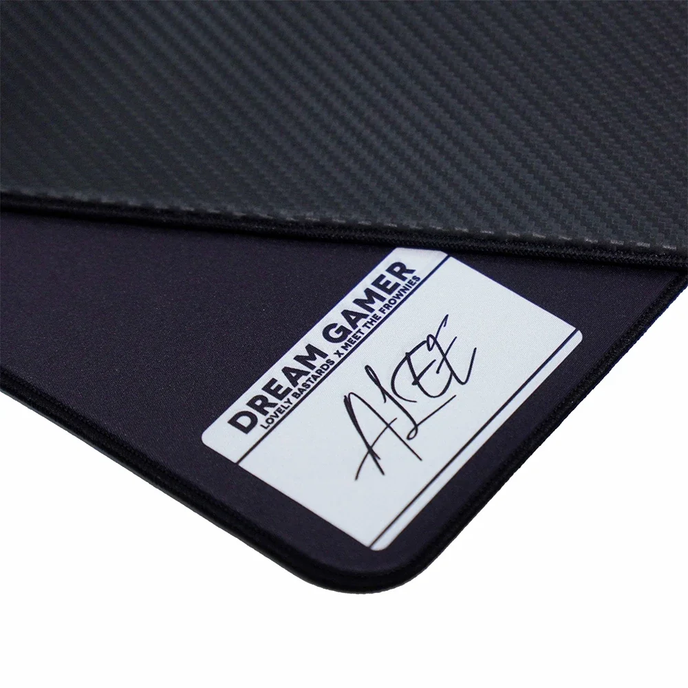 Karbon fiber desenli siyah oyuncu mouse pad'i ve üzerinde 'DREAM GAMER' yazısı ile imza bulunan beyaz bir etiket.