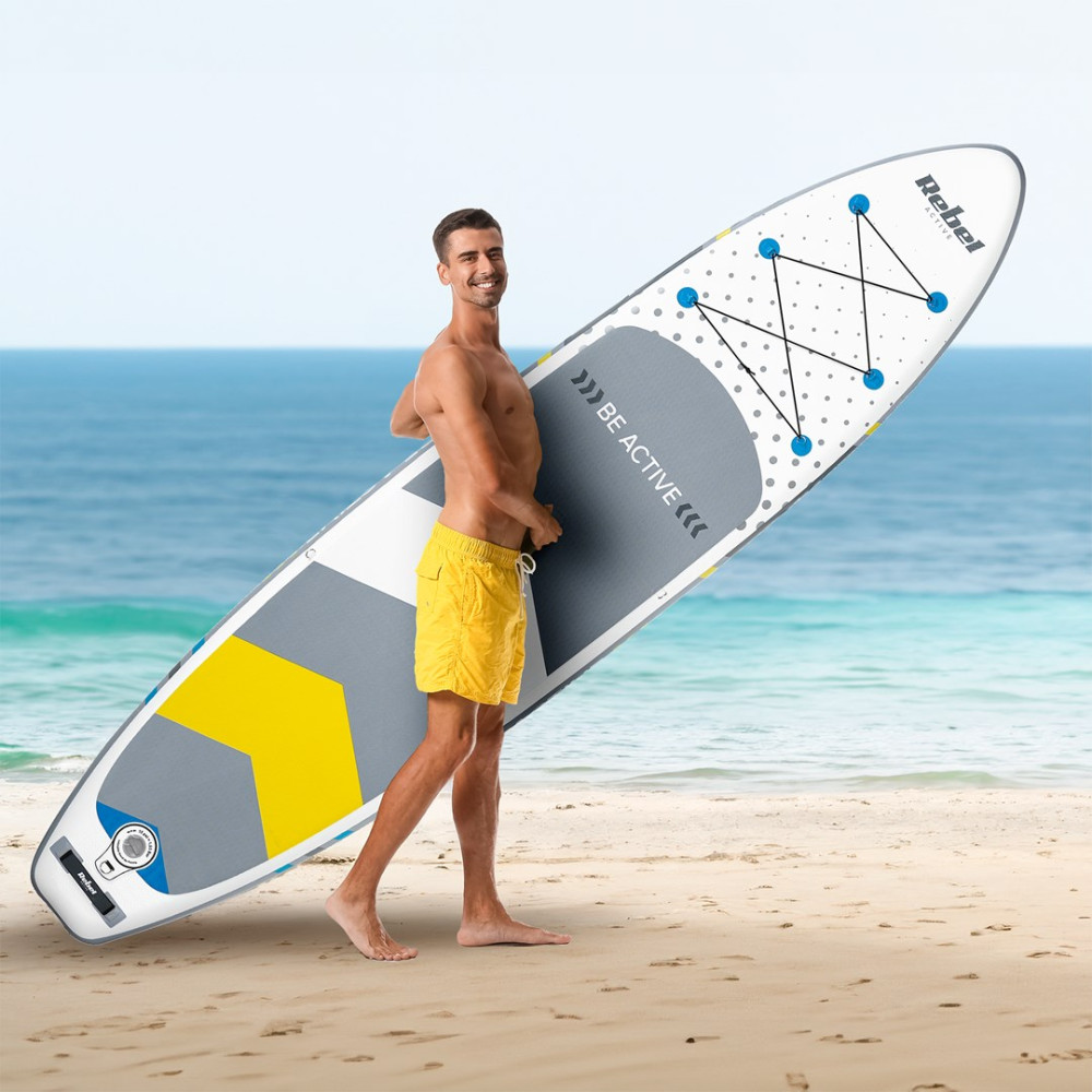 Un uomo sorridente a petto nudo con pantaloncini gialli tiene una tavola da SUP su una spiaggia soleggiata.