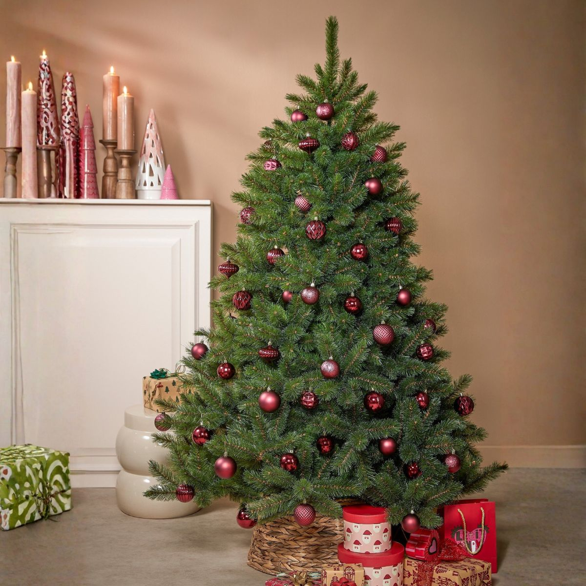 A decorated Christmas tree with red baubles stands in front of a wall, lit by candles on a mantelpiece.