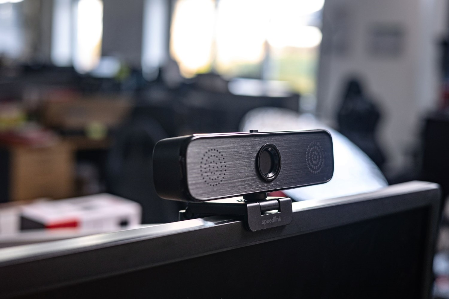 Eine schwarze Speedlink-Webcam ist auf einem Computermonitor montiert, im Hintergrund ist ein Büro zu sehen.