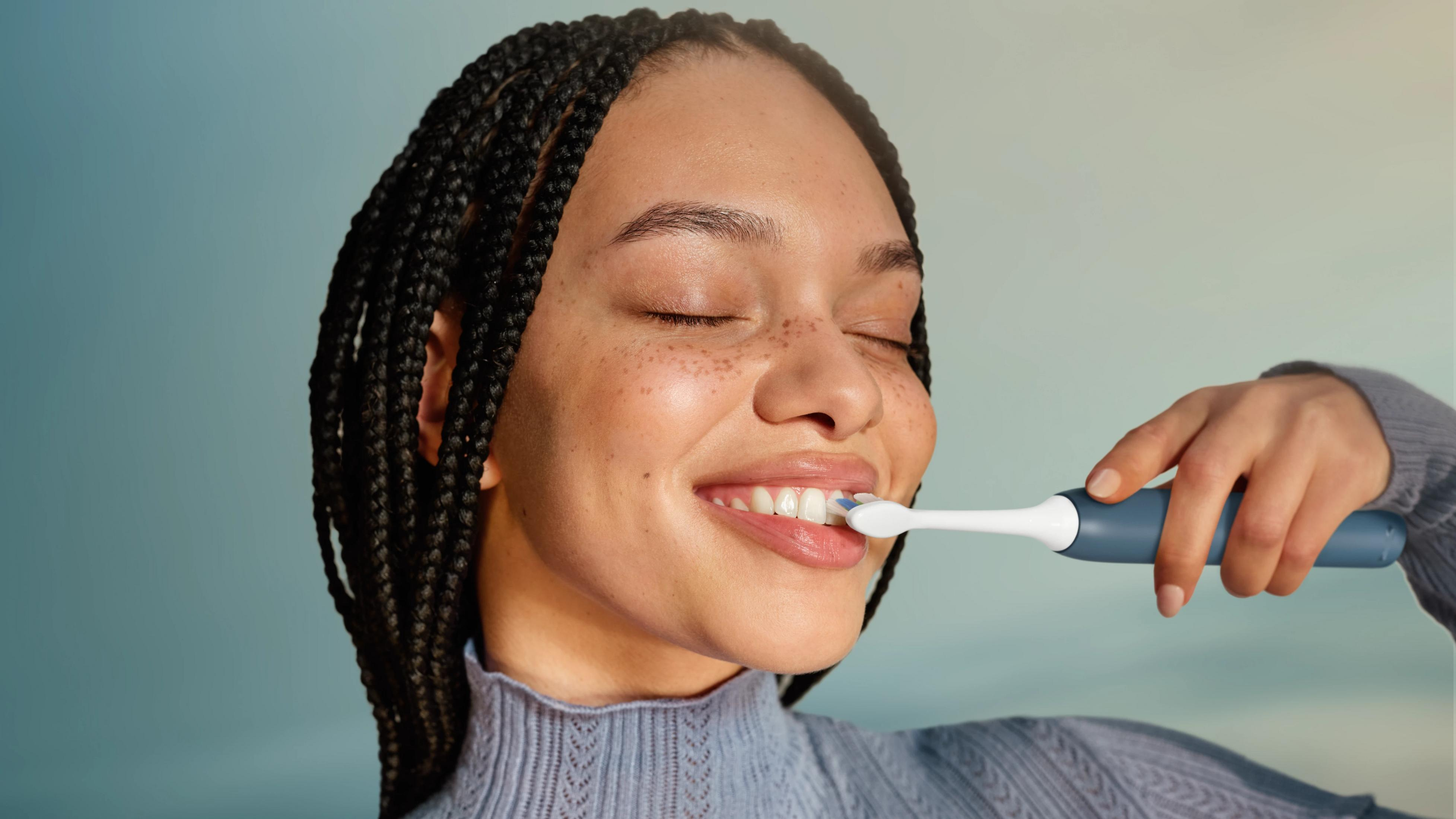 Una giovane donna con i capelli treccine sorride a occhi chiusi mentre si lava i denti con uno spazzolino elettrico.