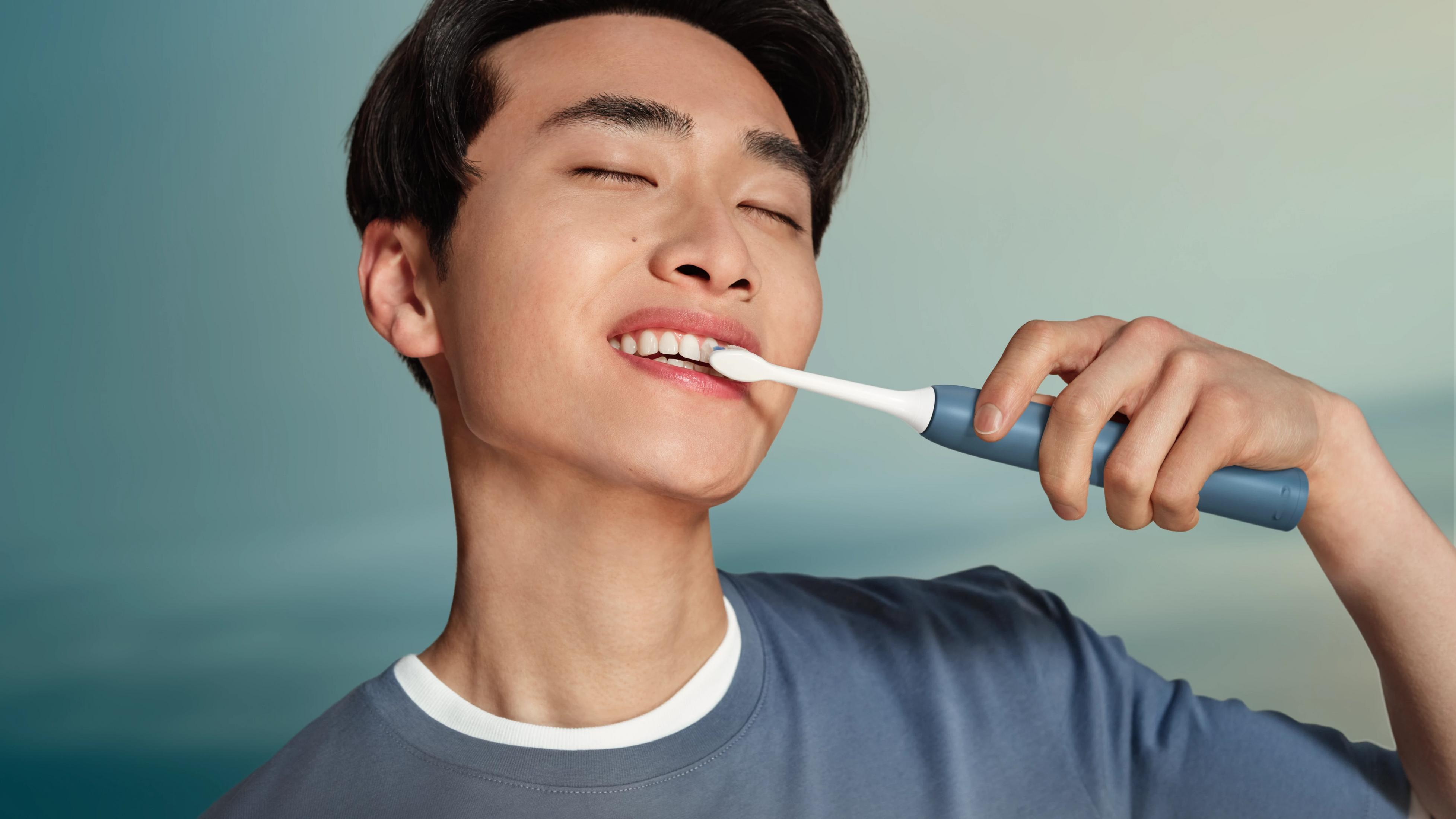 Un giovane sorride a occhi chiusi, lavandosi i denti con uno spazzolino elettrico blu.