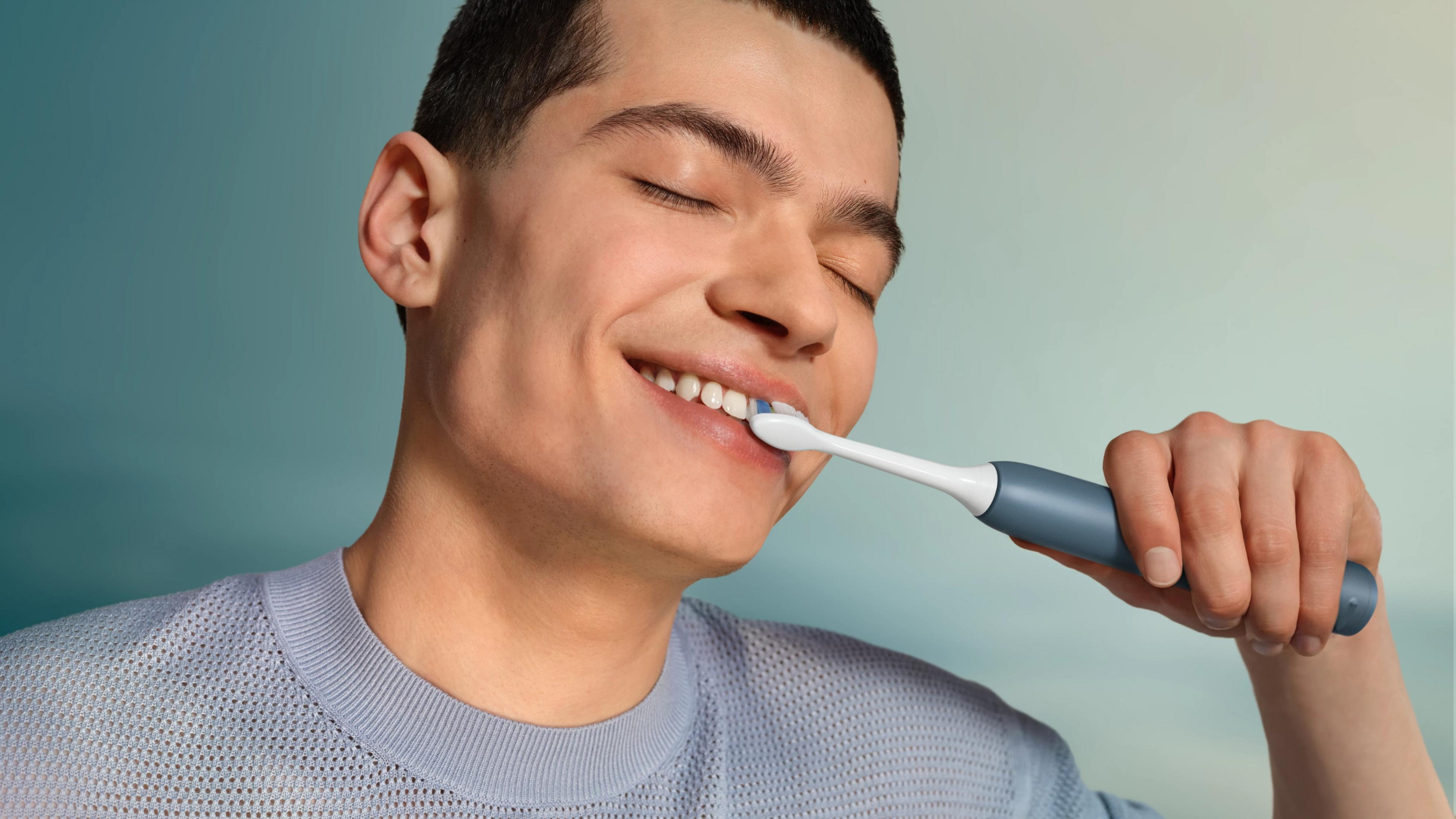 Un uomo sorridente con gli occhi chiusi si lava i denti con uno spazzolino elettrico su sfondo blu.