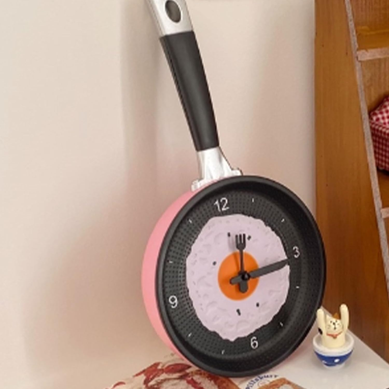 Eine bratpfannenförmige Wanduhr mit Spiegelei-Zifferblatt, Gabel- und Löffelzeigern, neben einer Katzenfigur.