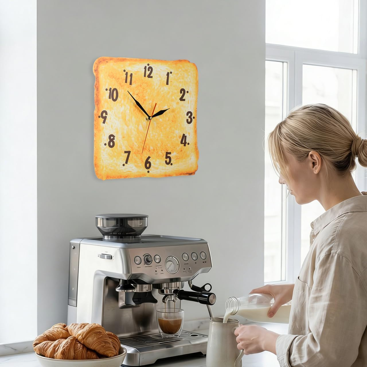 Eine blonde Frau gießt Milch in eine Tasse neben einer Espressomaschine und einer Toastbrot-Uhr an der Wand.