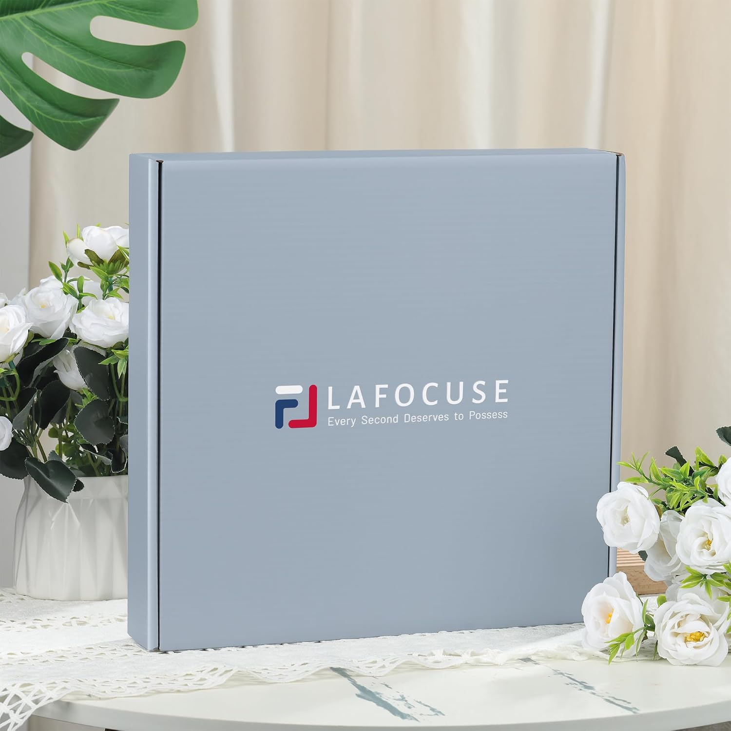 Eine graue La-Focus-Geschenkbox auf einem Tisch, flankiert von weißen Blumen und Vorhängen im Hintergrund.