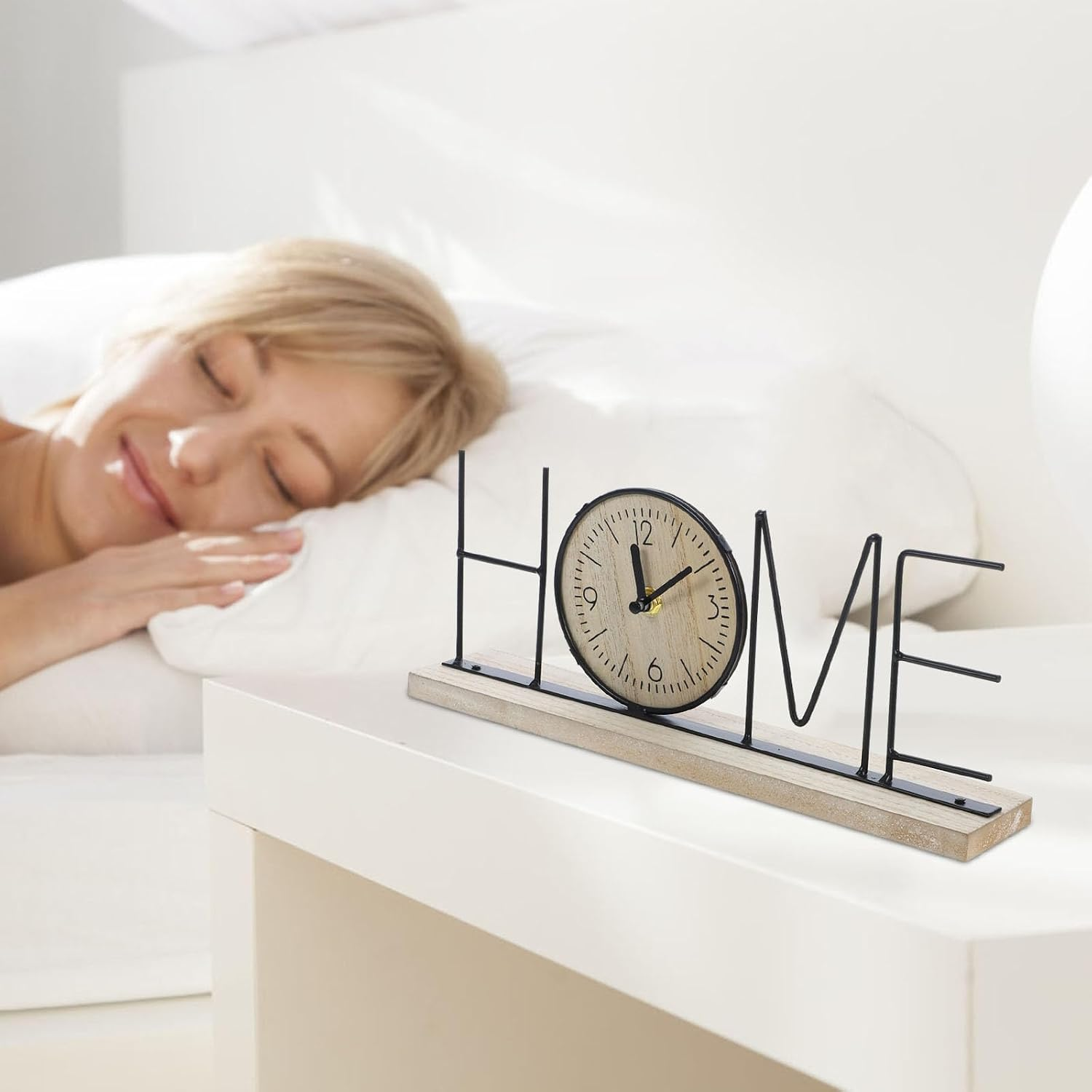 Ein hölzernes 'HOME'-Schild mit Uhr auf einem weißen Nachtschränkchen, im Hintergrund schläft eine Frau.