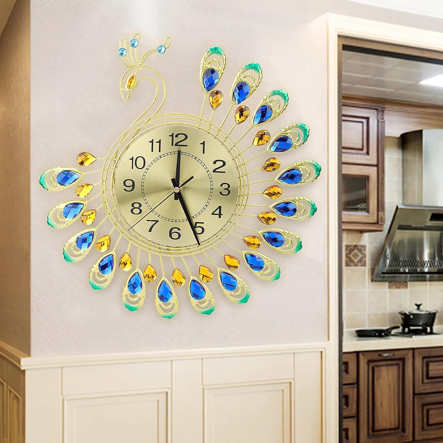 Eine goldene pfauenförmige Wanduhr mit blauen und gelben Kristallfedern hängt an einer hellen Wand.