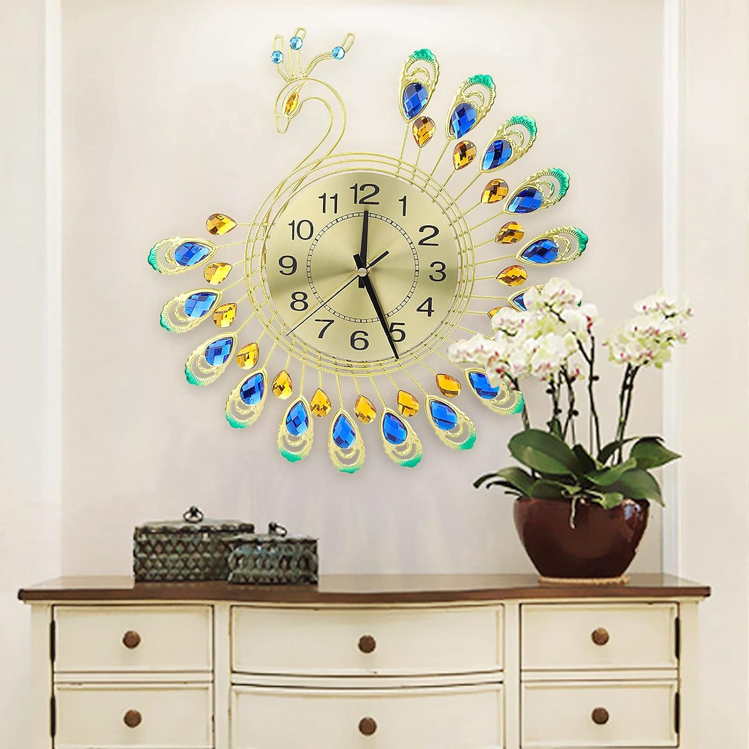 Eine goldene Pfauenwanduhr mit blau-goldenen Kristallen hängt über einer weißen Kommode mit Blumen.