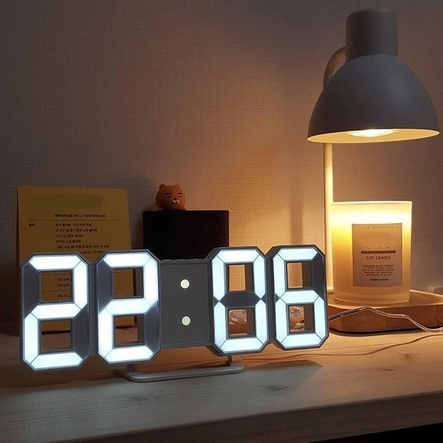 Eine weiße Digitaluhr zeigt 22:06 auf einem Holztisch, neben einer Lampe und einer Duftkerze.