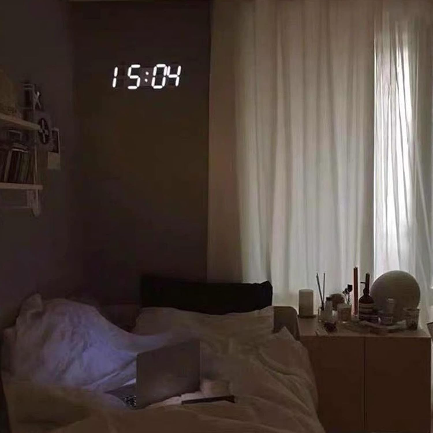 Ein gedimmtes Schlafzimmer mit einer Digitaluhr an der dunklen Wand, einem Bett mit Laptop und einem Nachttisch.