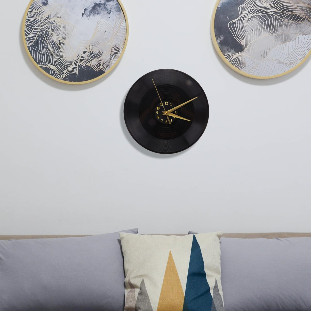 Eine Schallplattenuhr hängt an einer weißen Wand zwischen zwei runden Kunstwerken über einem Sofa mit Kissen.