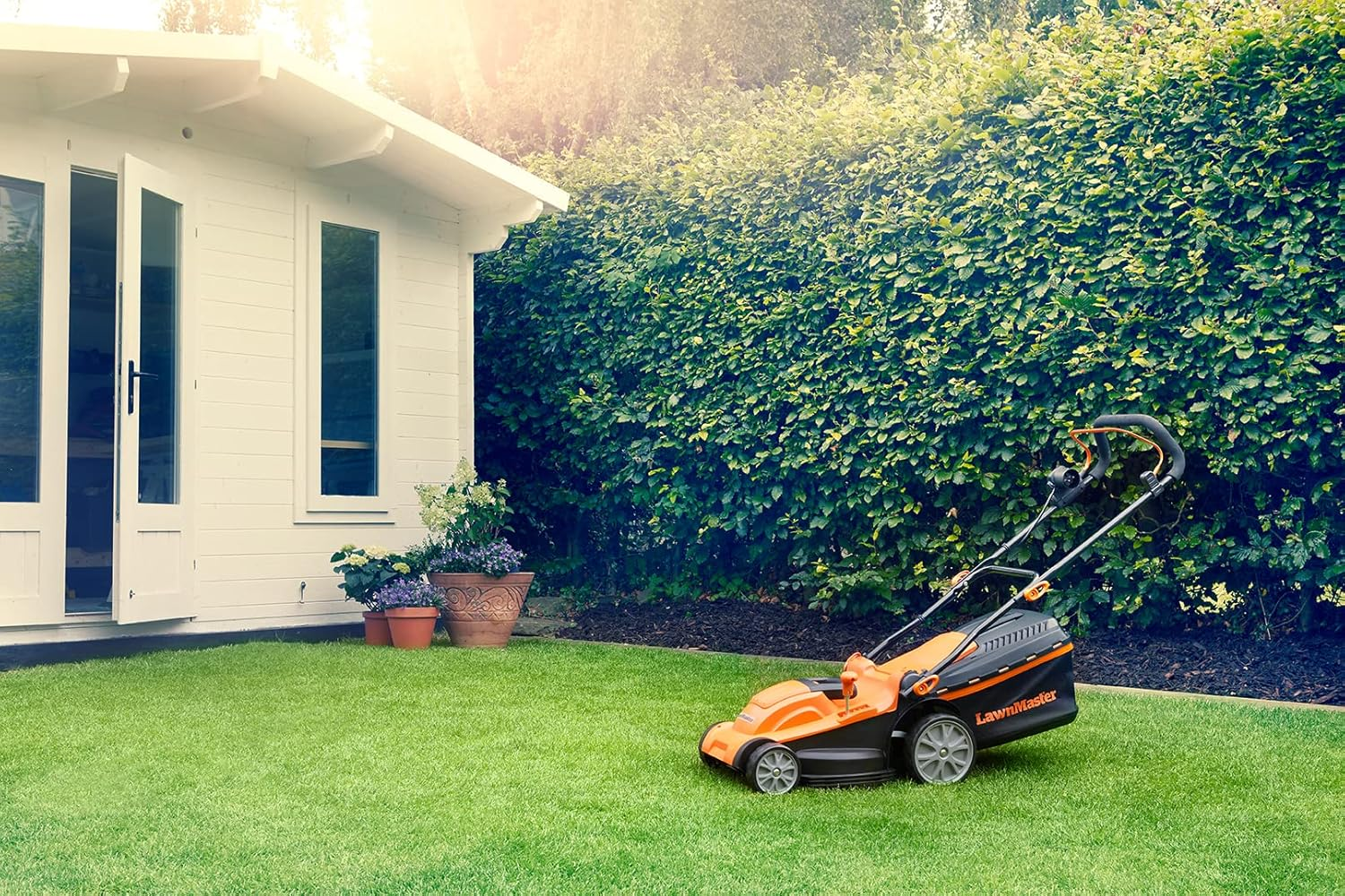 Ein orangefarbener LawnMaster-Rasenmäher steht auf grünem Gras vor einem weißen Gartenhaus und einer Hecke.