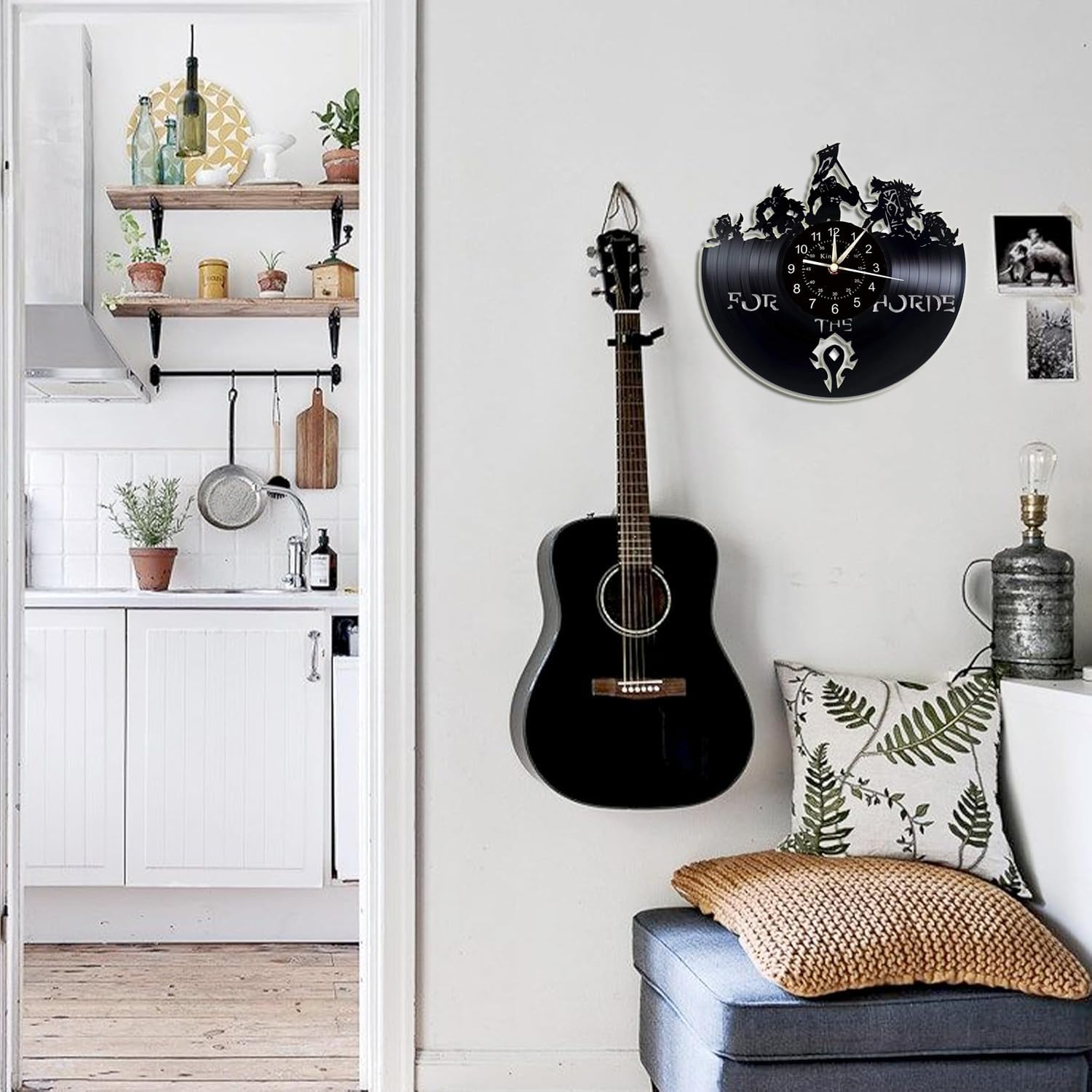 Eine schwarze Gitarre hängt an einer Wand neben einer Vinyl-Uhr und Kissen, im Hintergrund ist eine Küche sichtbar.