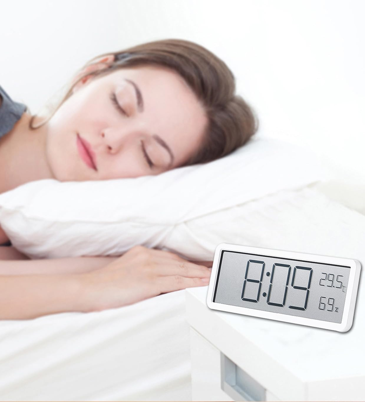 Eine junge Frau schläft friedlich im Bett, im Vordergrund steht ein digitaler Wecker auf einem Nachttisch.