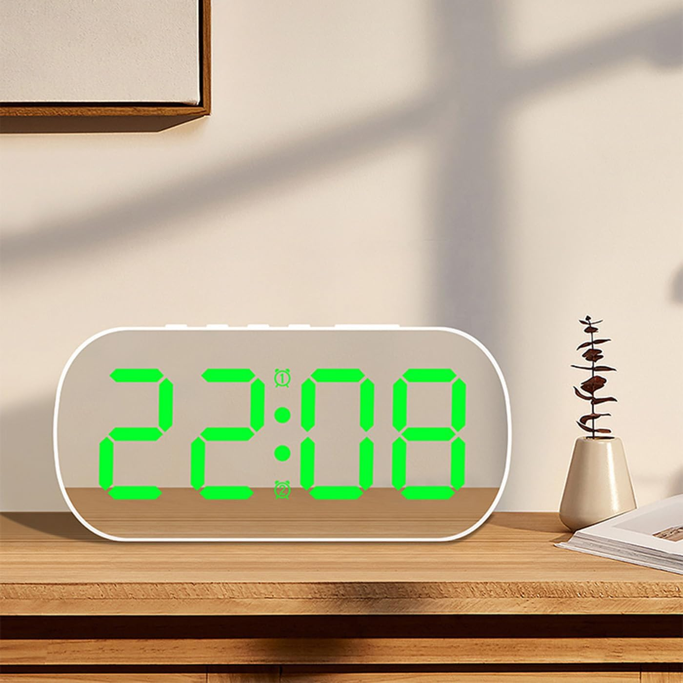 Ein weißer, ovaler Digitalwecker mit grüner LED-Anzeige steht auf einem Holztisch, daneben eine Vase mit Zweig.