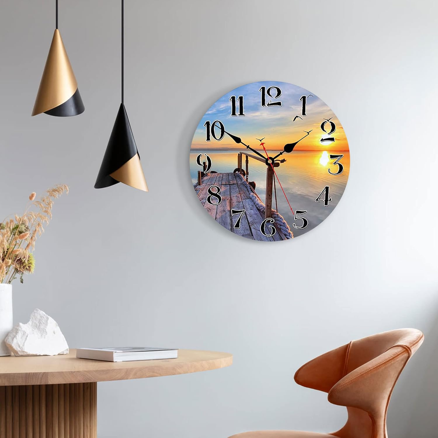Eine Wanduhr mit einem Steg-Motiv bei Sonnenuntergang hängt an einer grauen Wand neben Lampen.