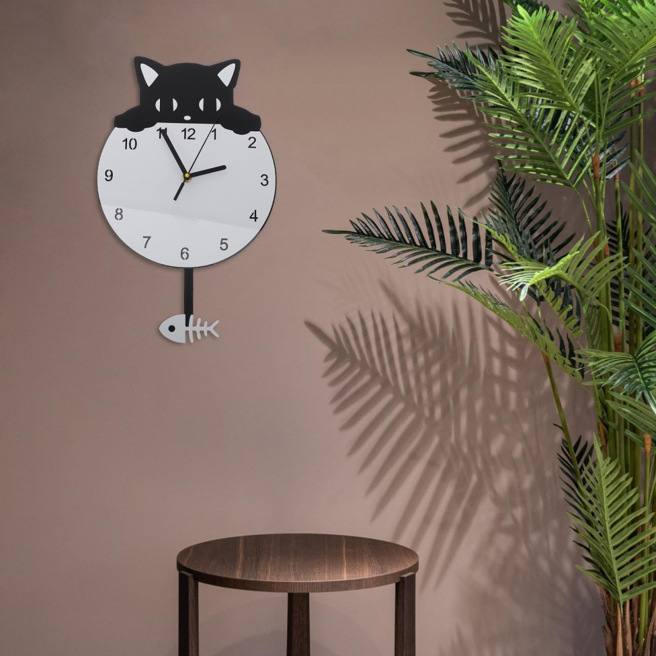 Schwarze Katzen-Wanduhr mit weißem Zifferblatt und einem Fisch-Pendel an einer Wand neben einer Pflanze.