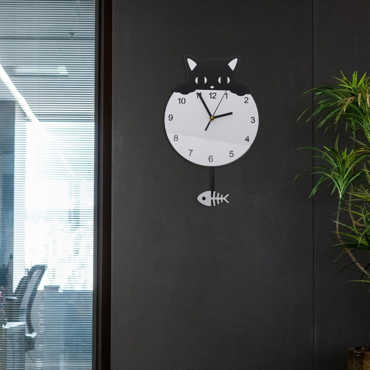 Eine schwarze Katzen-Wanduhr mit Pendel in Fischform hängt an einer dunklen Wand neben einer Pflanze.