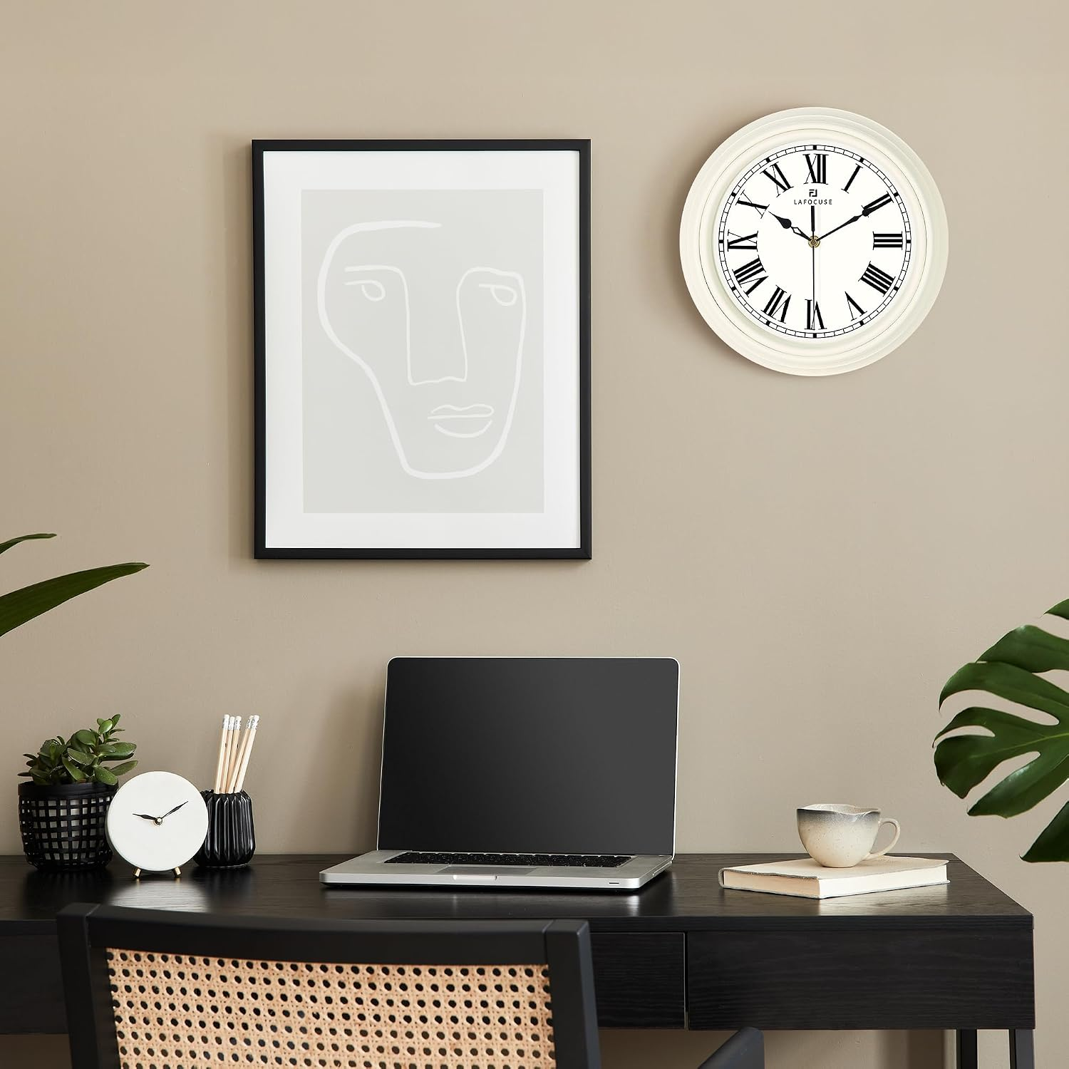 Ein ordentlicher schwarzer Schreibtisch mit Laptop, Wanduhr und gerahmtem Gesichtskunstwerk an einer beigen Wand.