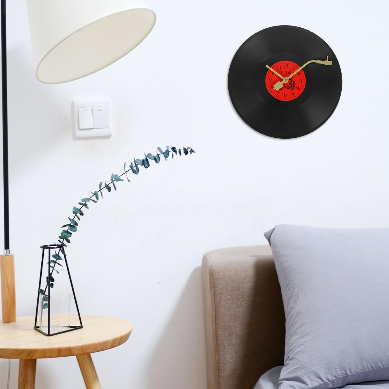 Eine Schallplatten-Wanduhr hängt nahe einer Lampe, einer Pflanze auf einem Beistelltisch und einem Bett mit grauem Kissen.