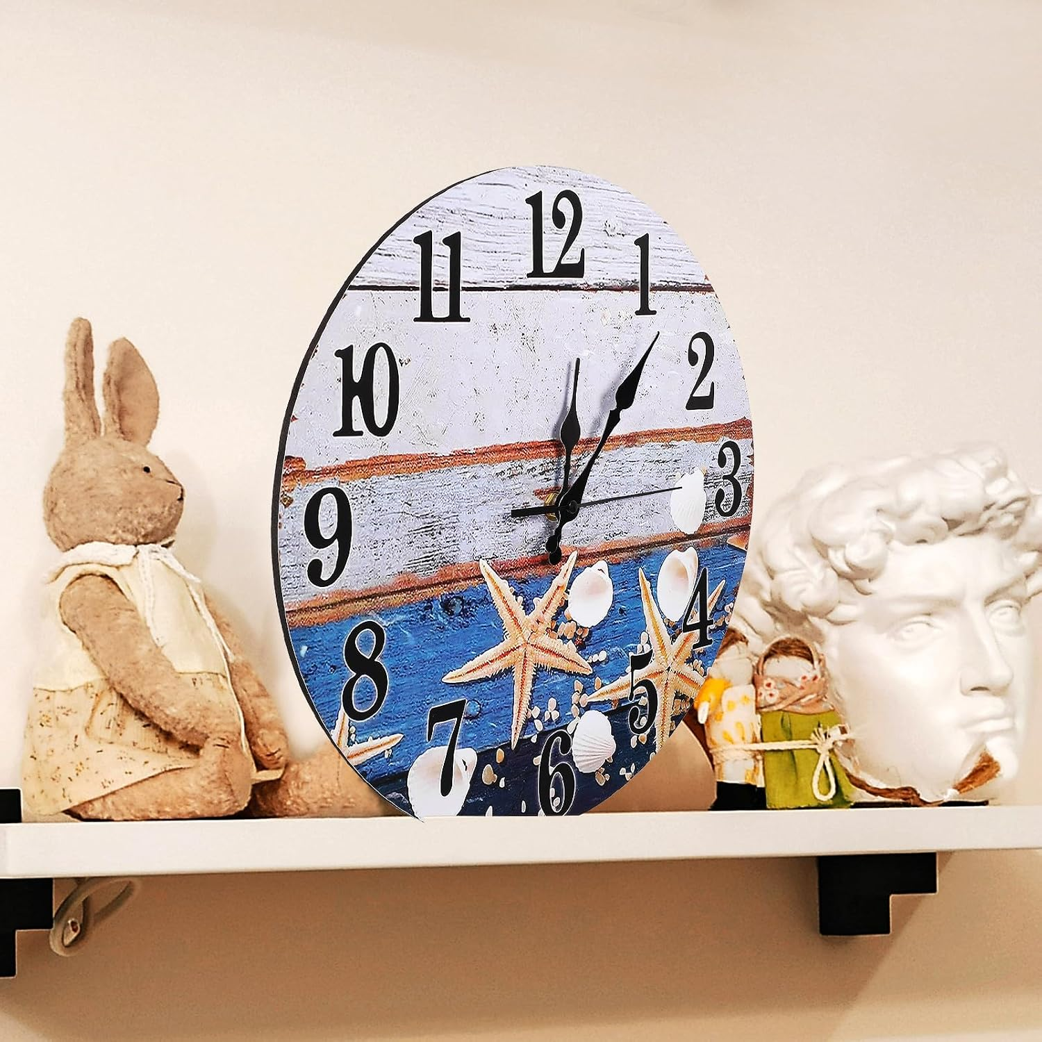 Eine Wanduhr mit Strandmotiv, ein Stoffhase und eine weiße Skulptur stehen auf einem weißen Regal vor einer Wand.