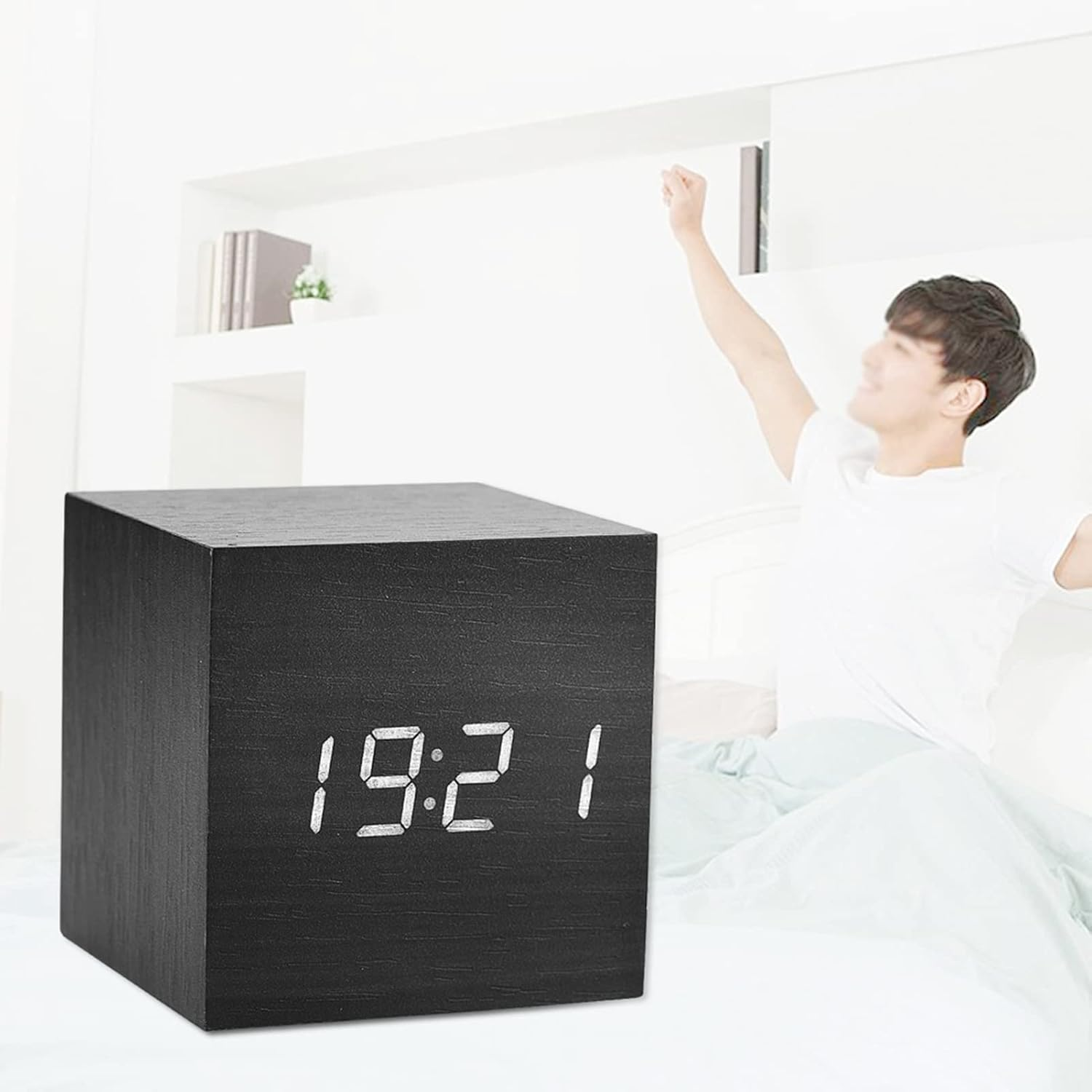 Ein schwarzer Holzwürfelwecker zeigt 19:21 Uhr, im Hintergrund streckt sich ein Mann im Bett.