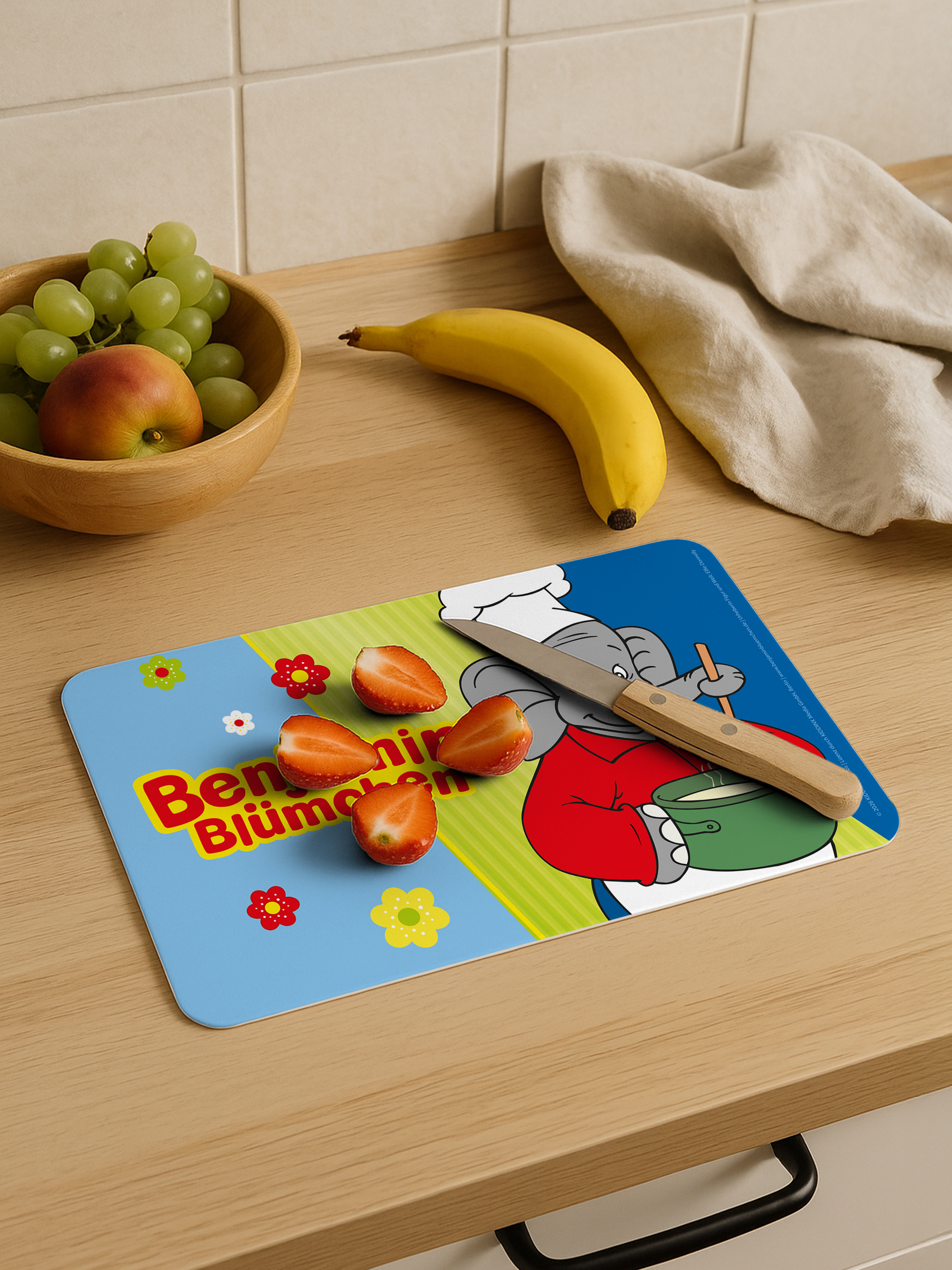 Ein Schneidebrett mit Elefantenmotiv, Erdbeeren und Messer auf Holz-Arbeitsplatte mit Früchten und Tuch.