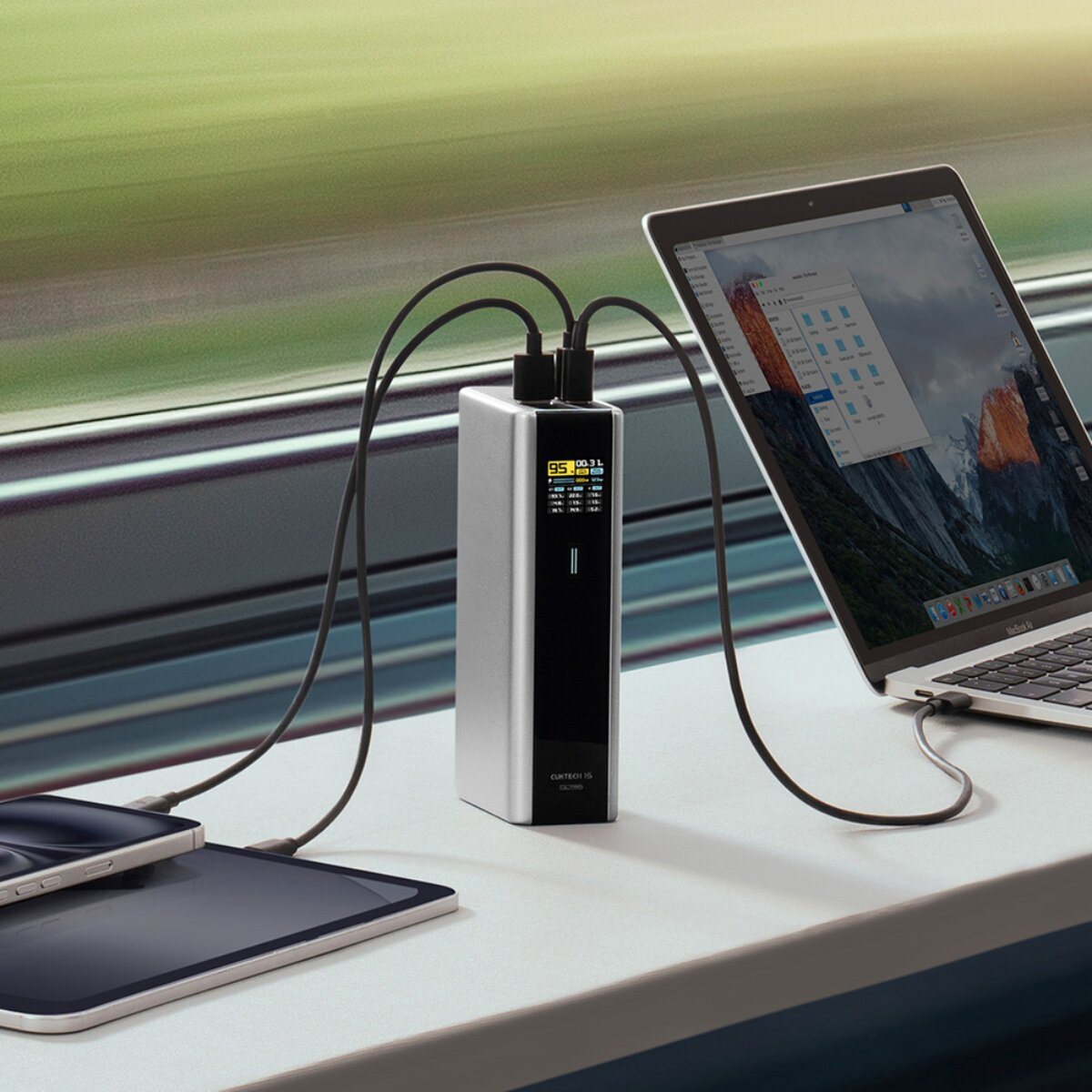 Eine silberne Powerbank mit Kabeln lädt Laptop, Tablet und Smartphone auf einem Zugtisch.