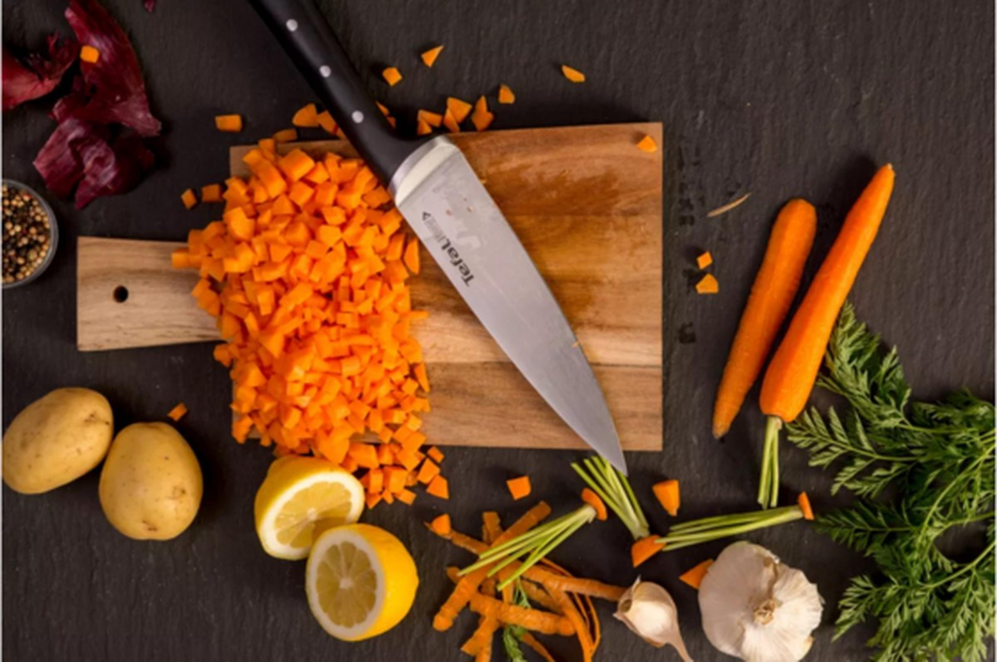 Un coltello da chef su un tagliere con carote tritate, carote intere, patate, limoni e aglio su una superficie di pietra scura.
