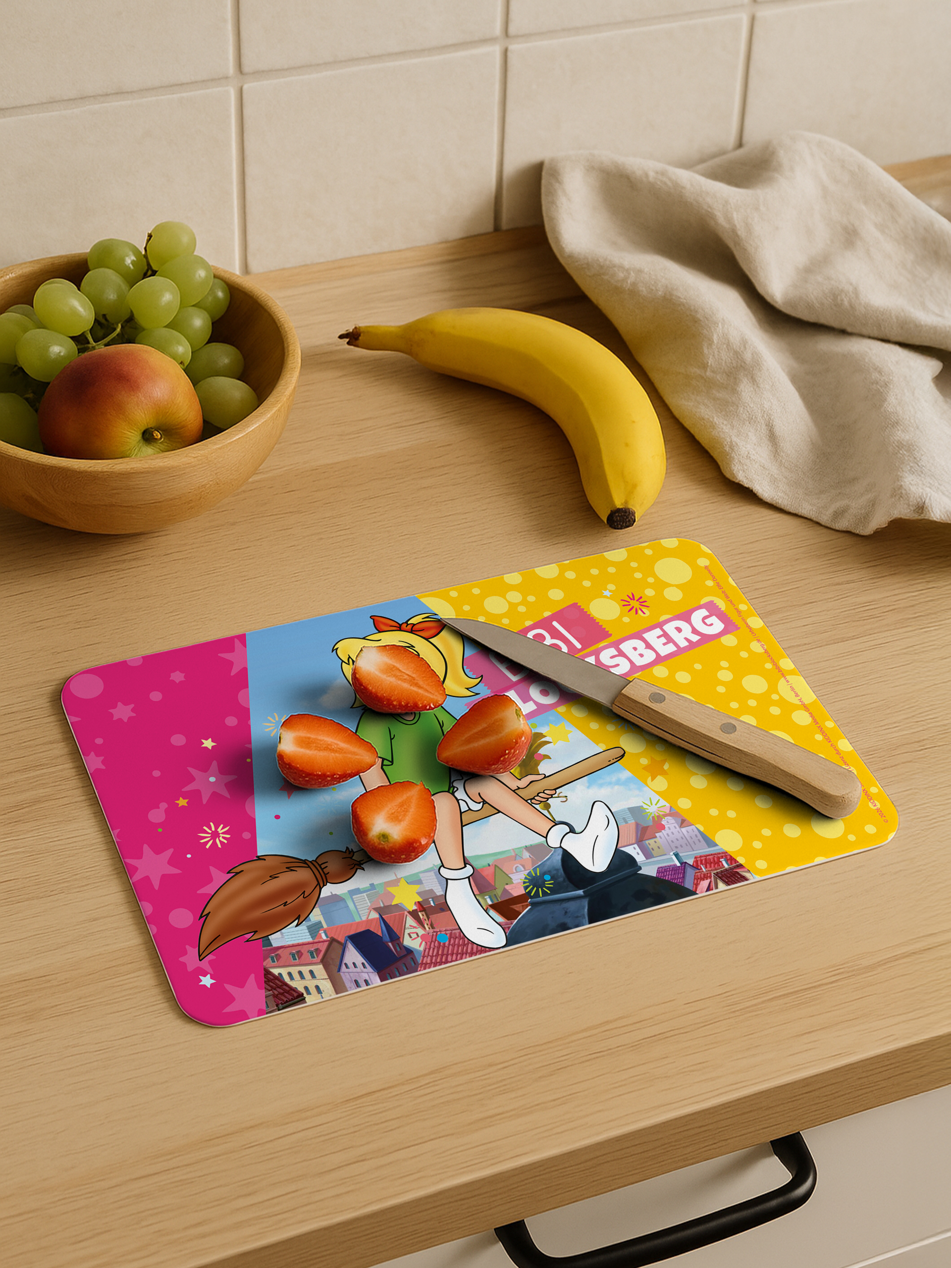Geschnittene Erdbeeren und ein Messer auf einem bunten Schneidebrett neben einer Banane.