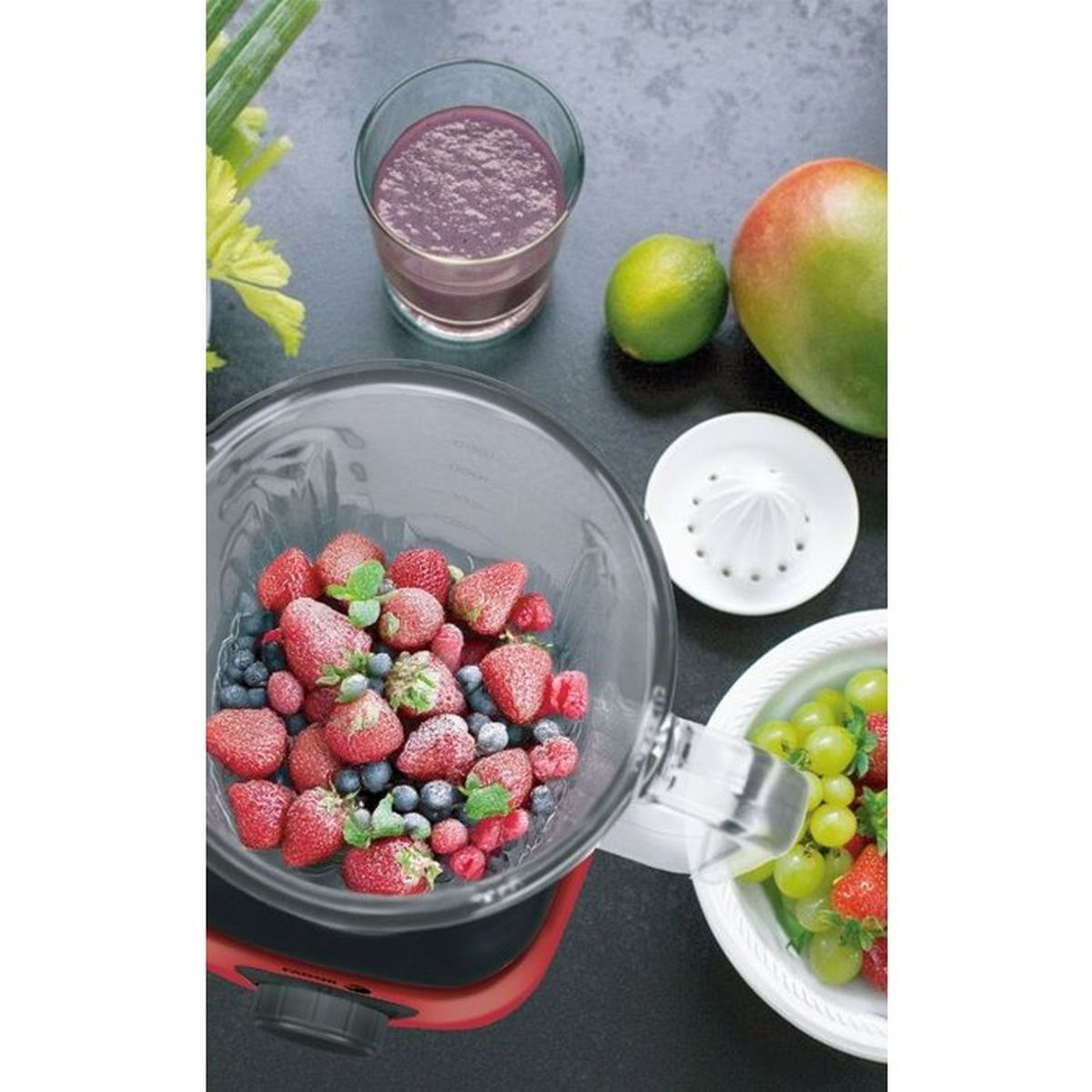 Un frullatore pieno di fragole e mirtilli, circondato da frutta, succo e uno spremiagrumi.