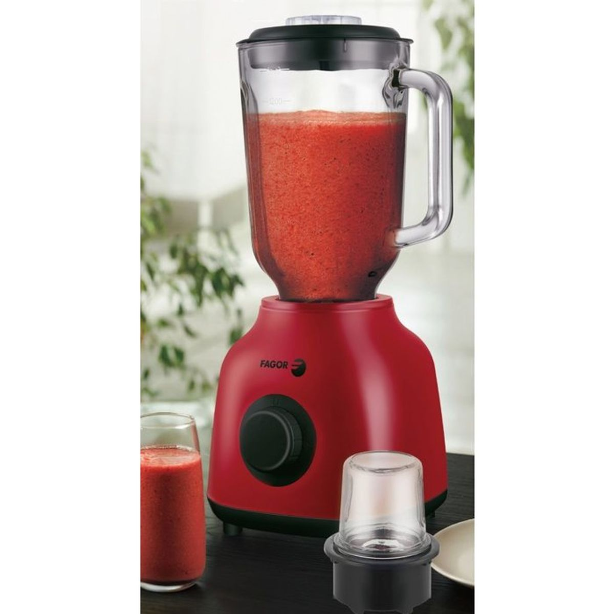 Un frullatore rosso pieno di frullato rosso su un tavolo, con un bicchiere di frullato e un accessorio macinino vicino.