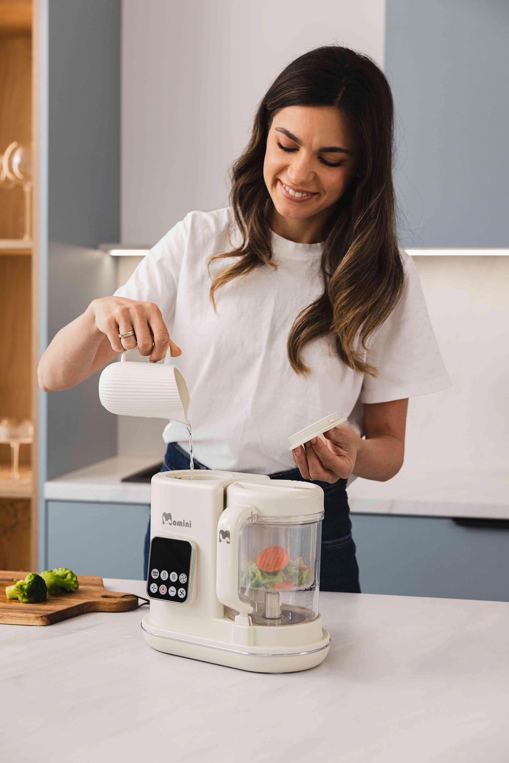 Una donna sorridente versa acqua in un elettrodomestico bianco su un bancone, con broccoli.
