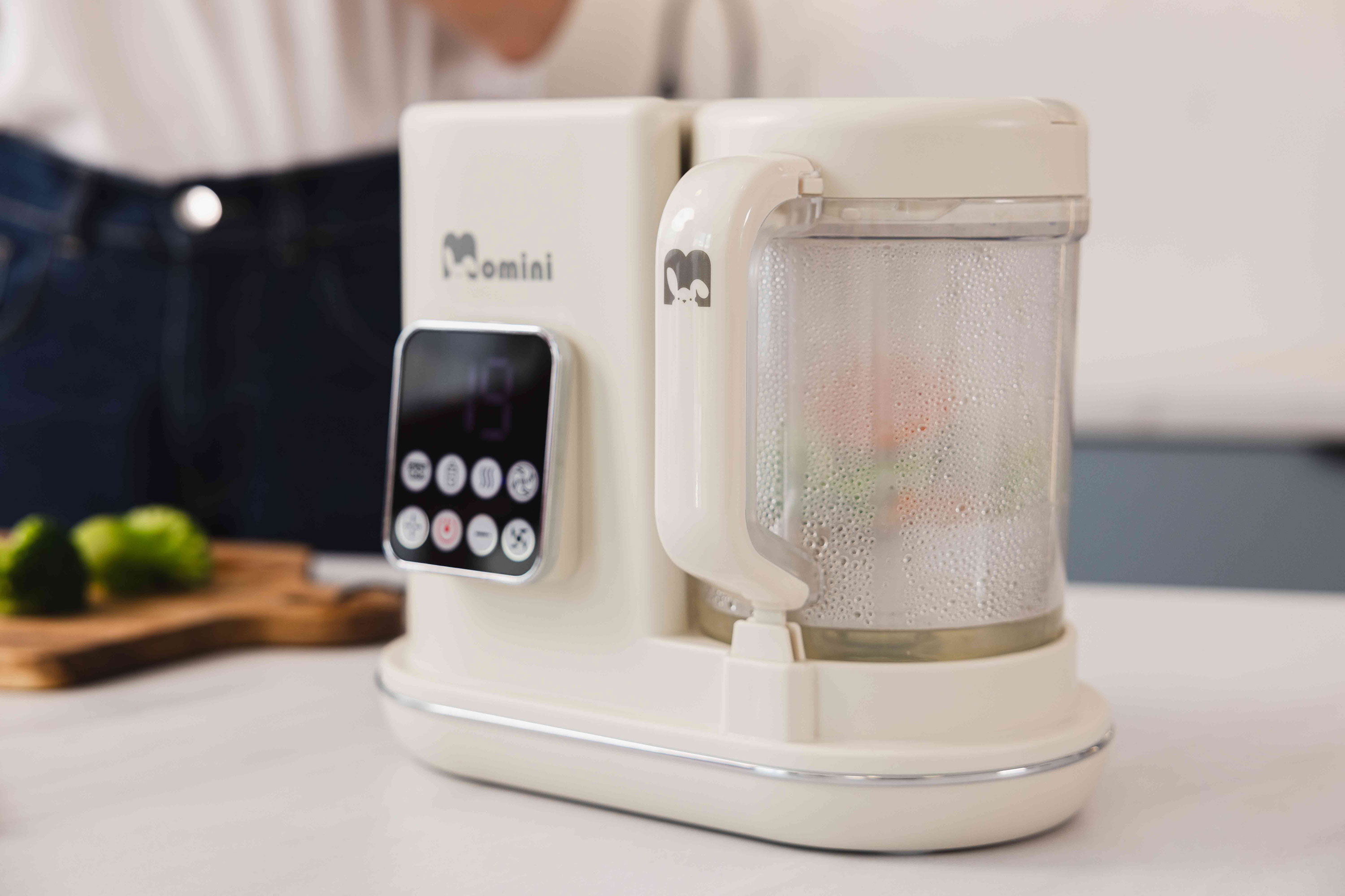 Un robot da cucina bianco Momini si trova su un bancone chiaro in cucina, con un boccale per la cottura a vapore con verdure.