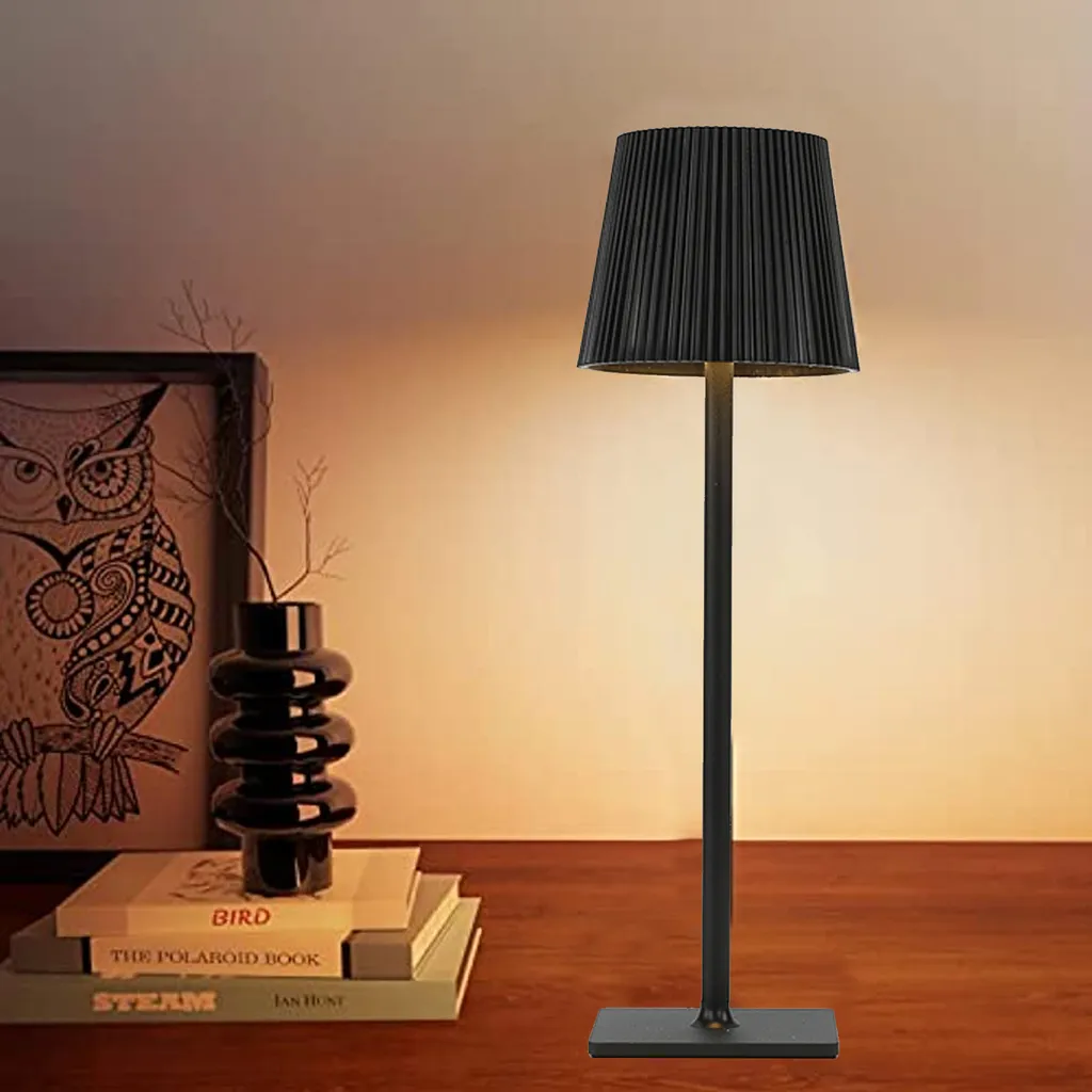 Eine schwarze moderne Tischlampe steht auf einem Holzschreibtisch neben gestapelten Büchern, einer schwarzen Vase und einem Eulenbild.