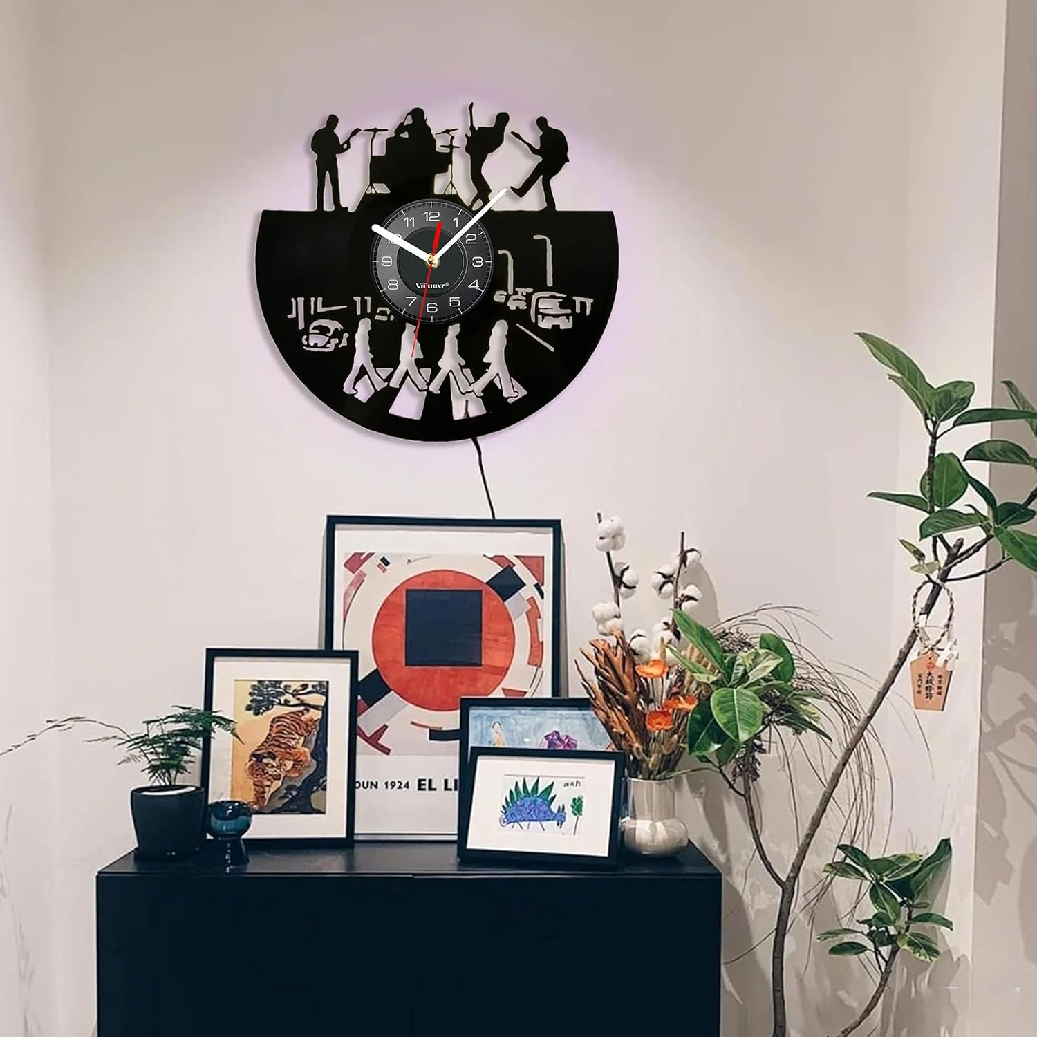 Eine schwarze Wanduhr in Vinyl-Form mit Bandsilhouetten hängt über einem dunklen Schrank mit gerahmter Kunst.