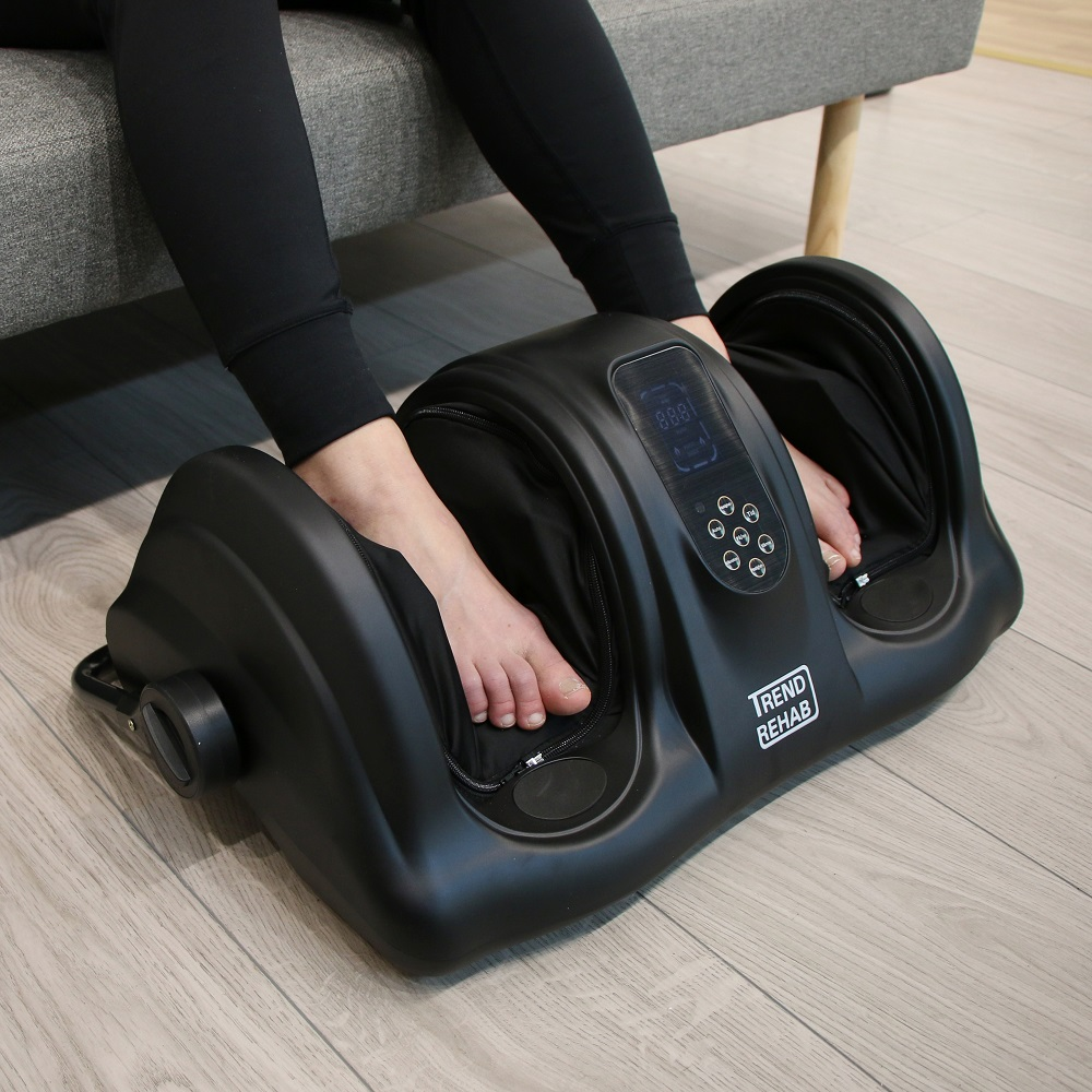 Eine Person sitzt auf einem Sofa und nutzt ein schwarzes Trend-Rehab-Fußmassagegerät auf einem Holzboden.